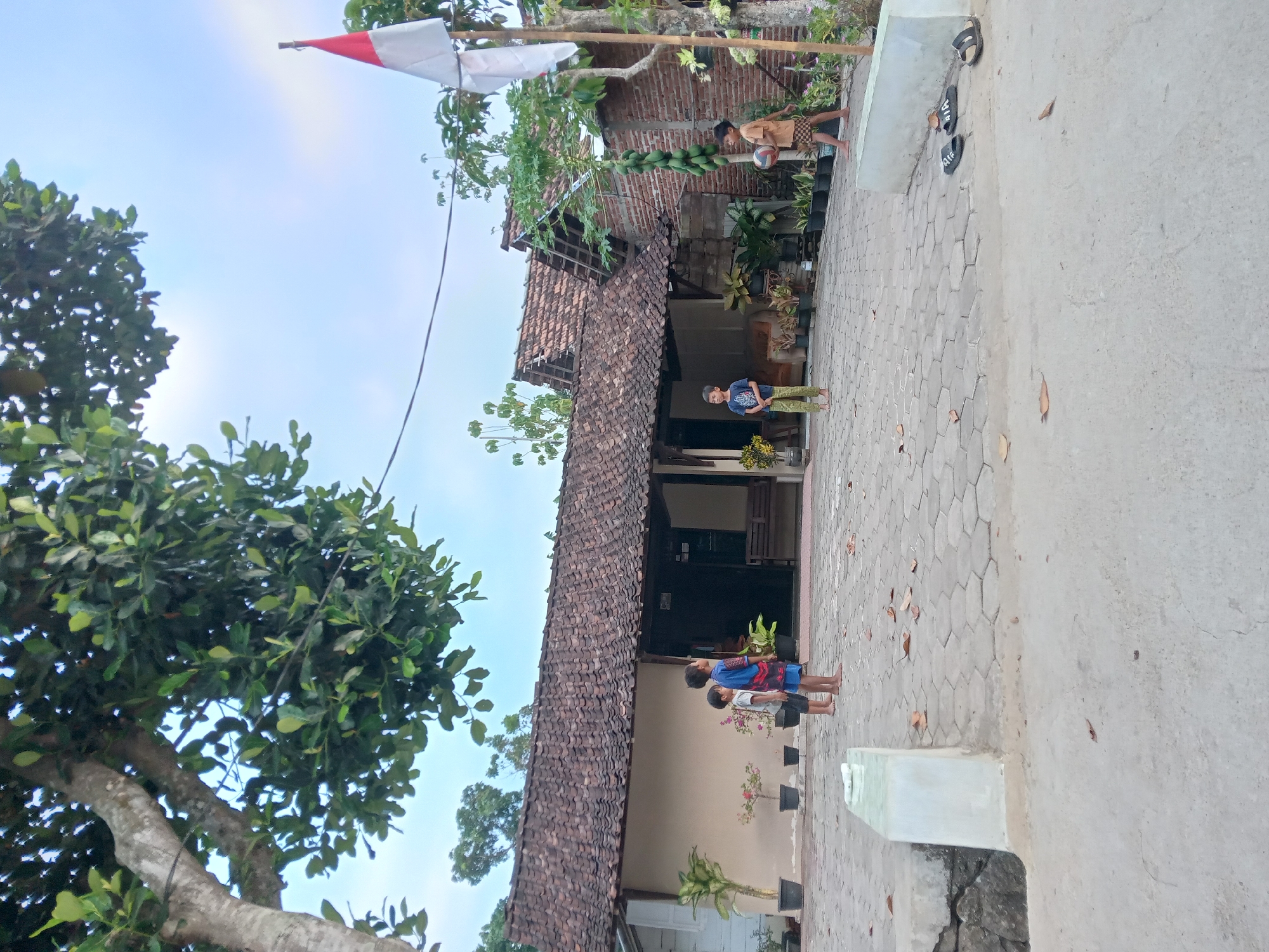 In Kedunggudel, Indonesia, a residential scene unfolds under a hazy sky. A traditional Indonesian house with a tiled roof, featuring a prominent front entrance and a covered porch, forms the backdrop. In front of the house, a paved courtyard with hexagonal tiles serves as the primary area of activity. Several young boys are present, engaged in various activities. Two boys stand near the house, one in a blue shirt and patterned shorts, the other in a blue t-shirt and green pants. Further to the right, another boy in a light brown t-shirt and patterned shorts holds a basketball. A small Indonesian flag flies from a pole near the right edge of the frame. Lush green trees, one with a substantial trunk and canopy, partially obscure the sky. The environment appears to be a rural or semi-rural residential area.