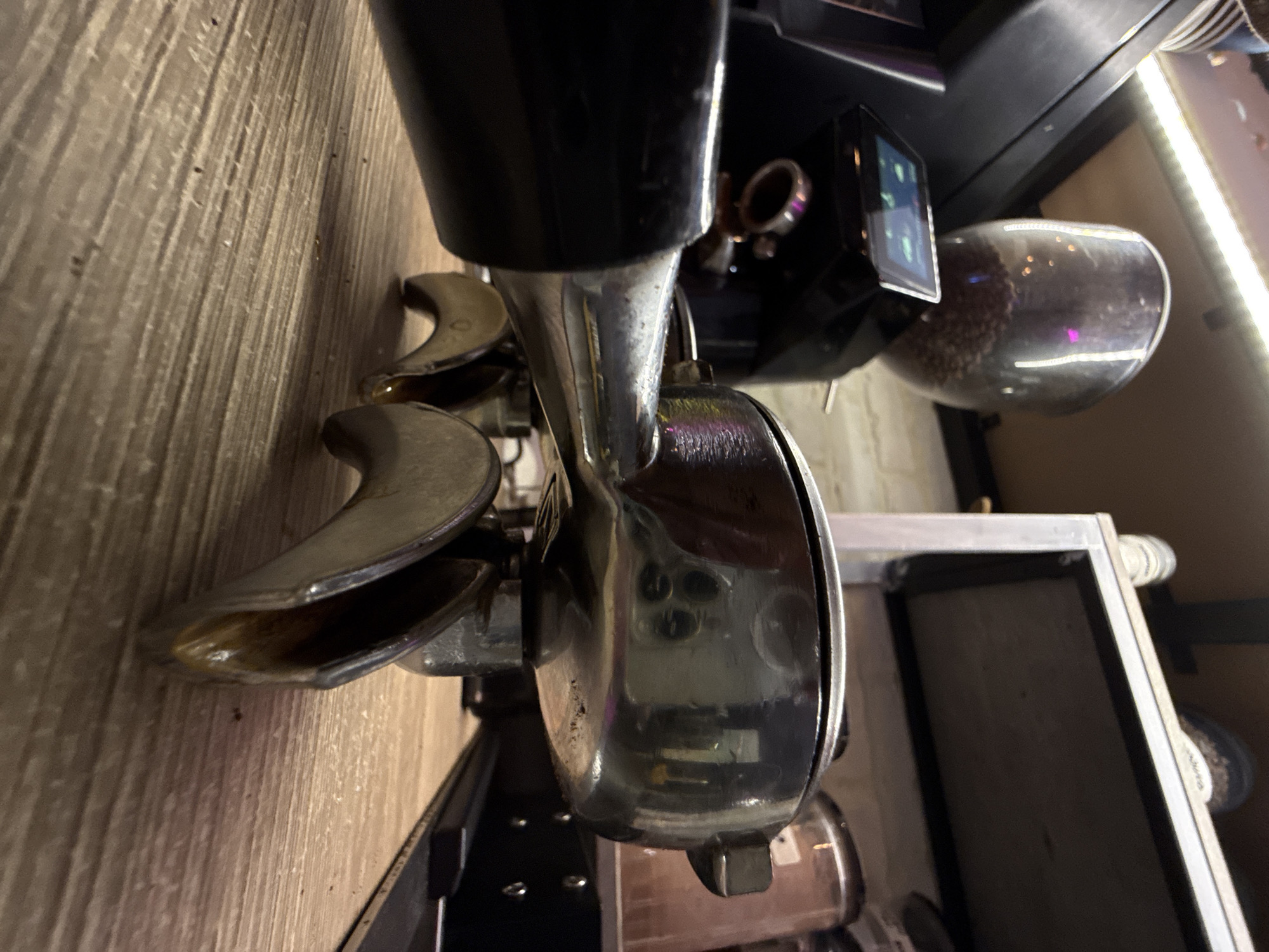 The image presents a close-up view of a coffee machine, with a prominent, shiny metal grinding component in the foreground. The machine's spout is visible, with remnants of coffee grounds within. Above the grinding mechanism, a dark-colored, possibly black, body of the machine is present, featuring a small display screen. To the right of the machine body is a glass container filled with coffee beans. The environment appears to be a countertop surface with a wood-grain pattern. To the right, a partial view of a white shelving unit with a dark interior is present. Based on the prompt mentioning the location of Dolgoprudny, Russia, it can be inferred that this scene takes place in a café or kitchen setting within that city.