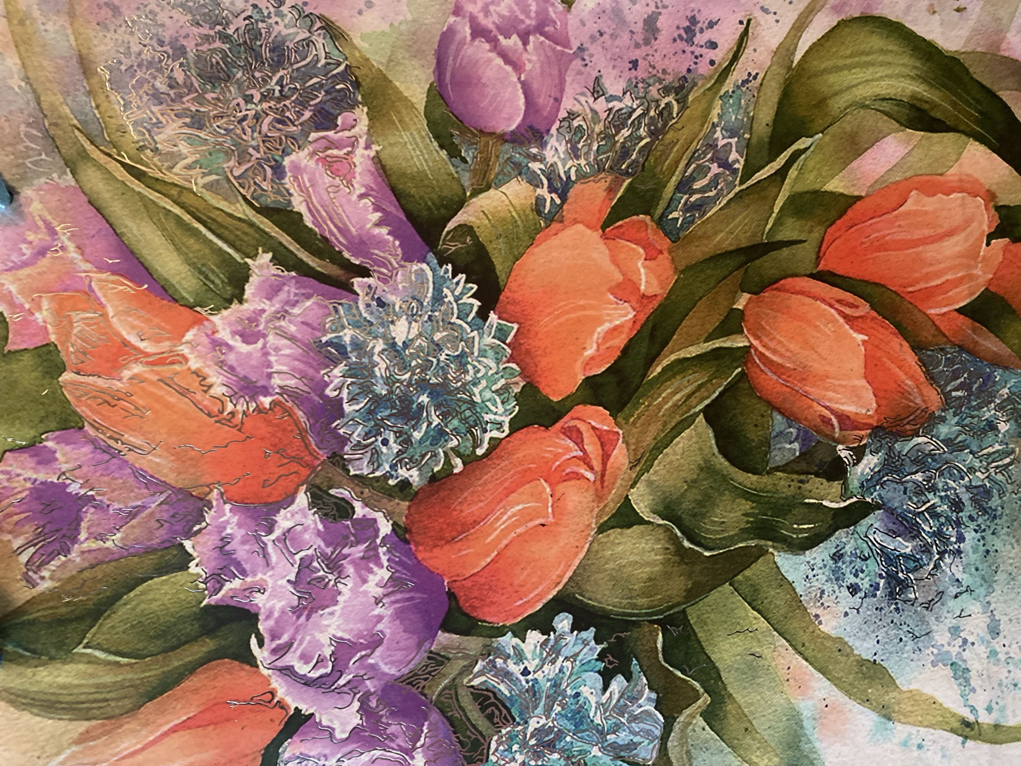 This is a close-up watercolor painting of a vibrant bouquet of flowers. The arrangement features a mix of orange tulips, purple hydrangeas, and some blue, spiky flowers, interspersed with lush green leaves. The paint application is loose and expressive, with visible brushstrokes and splatters, giving the artwork a dynamic and textured feel. The overall impression is one of abundant, colorful floral beauty. The painting is not located in any specific city; it is an artwork.