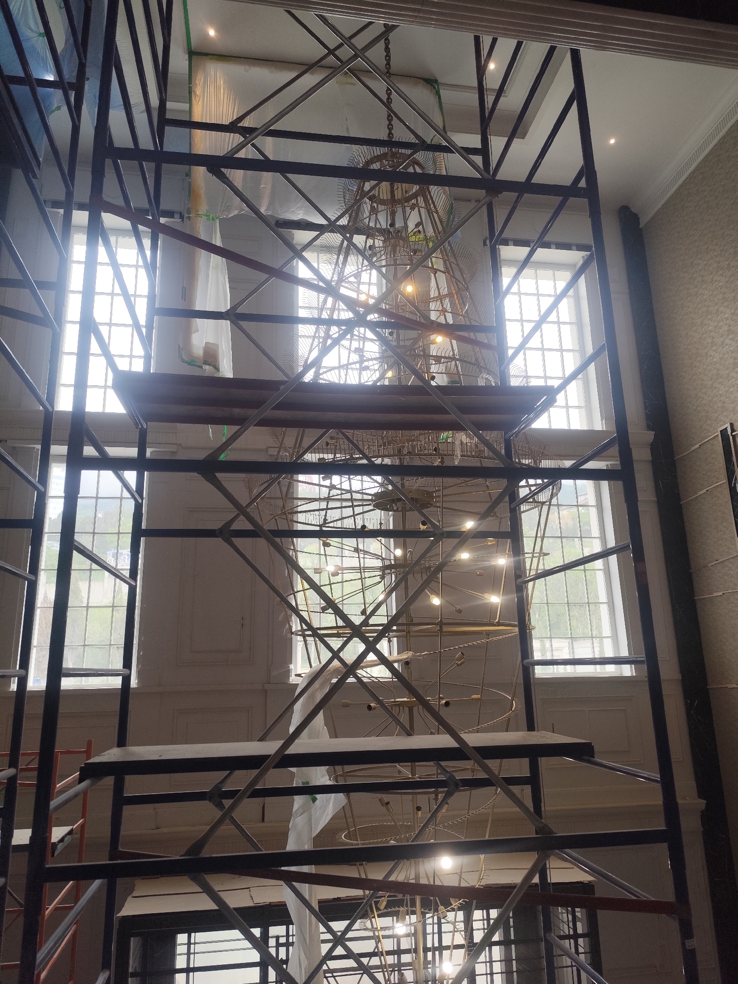 The image shows a tall, multi-tiered scaffolding structure dominating the foreground. A large, ornate chandelier or light fixture hangs partially obscured within the scaffolding. The backdrop consists of tall windows with a grid pattern, suggesting a grand interior space. The walls are painted white and there is decorative molding above the windows. A tall column with grey patterned wall. The image location and city are unknown.