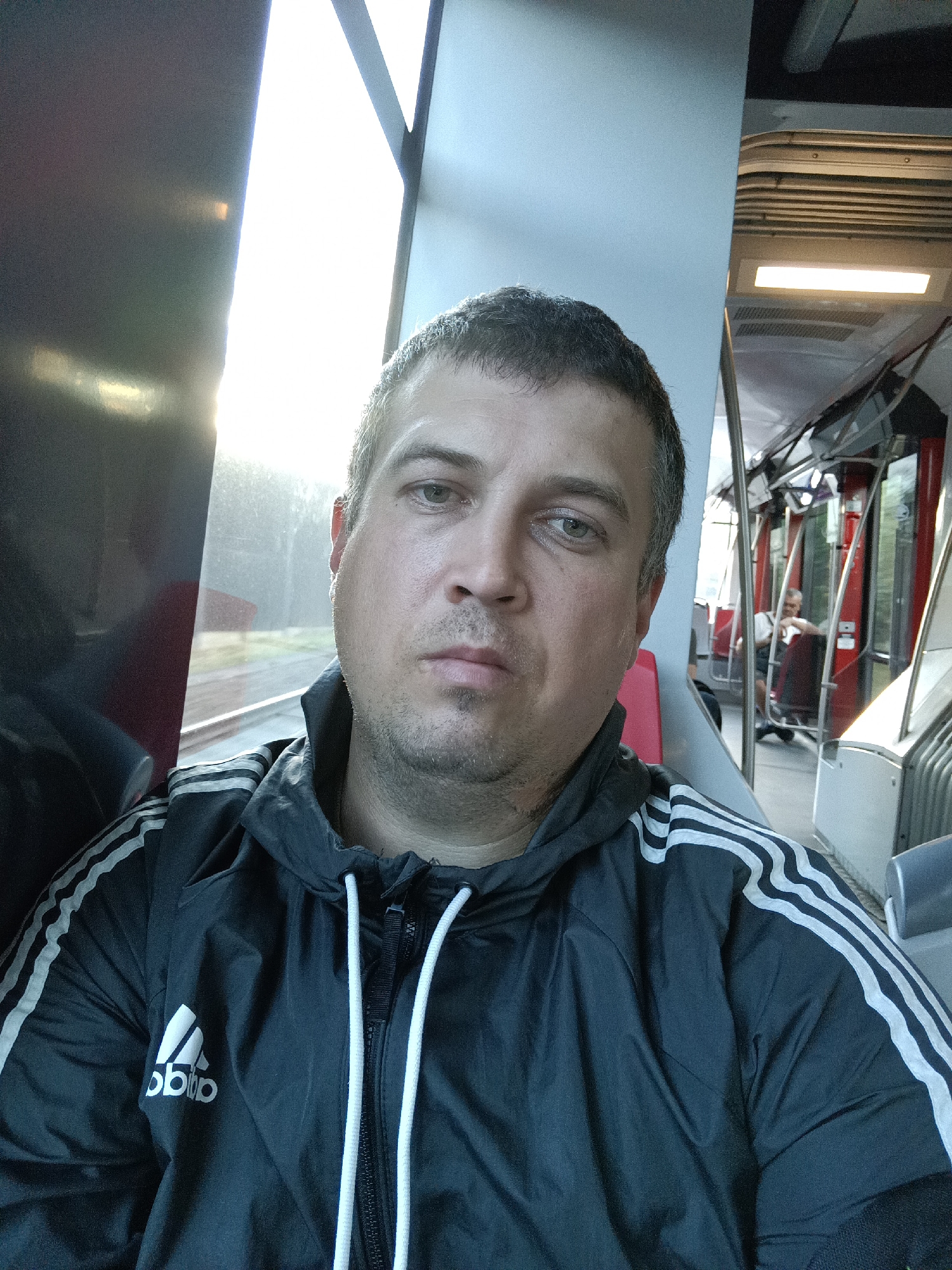 A mid-shot selfie captures a Caucasian man in his late 30s or early 40s, looking directly at the camera. He has short, dark hair and is wearing a black Adidas tracksuit jacket with white stripes on the sleeves. The man is seated inside a tram, with the interior of the tram visible behind him, including red seats and handrails. Sunlight streams in from a window to his left, creating a bright glare. The man's expression is neutral. In the background, another passenger is visible further down the tram. The setting appears to be a public transportation vehicle in Prague, Czechia.