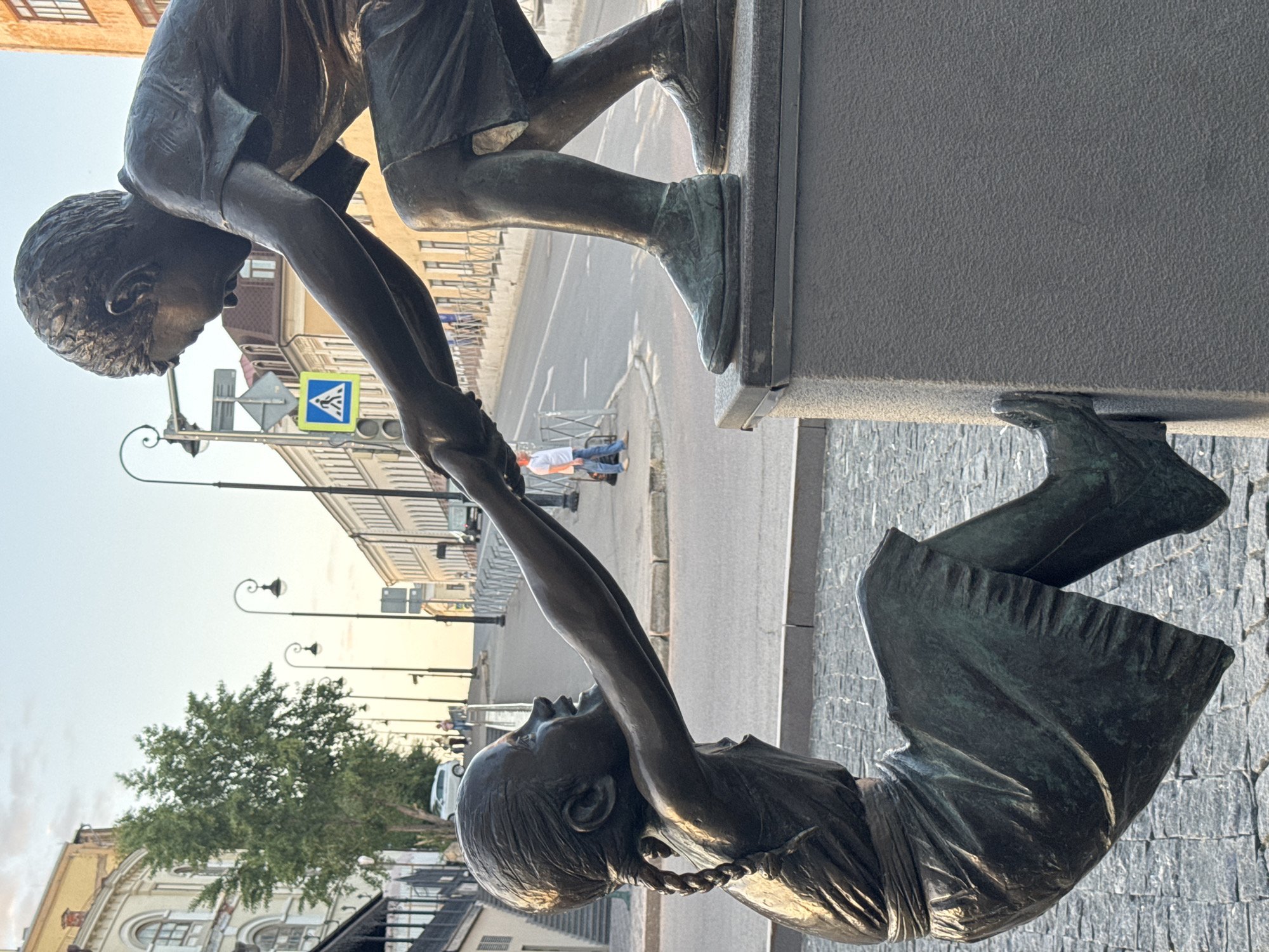 The image depicts a bronze statue in Kazan, Russia, titled "Touch". The statue shows a boy reaching down to help a girl who is below him, creating a visual of friendship and assistance. The boy is standing on a raised platform, while the girl is reaching up to him. The background includes a city street with buildings, streetlights, a pedestrian crossing sign, and people walking.
