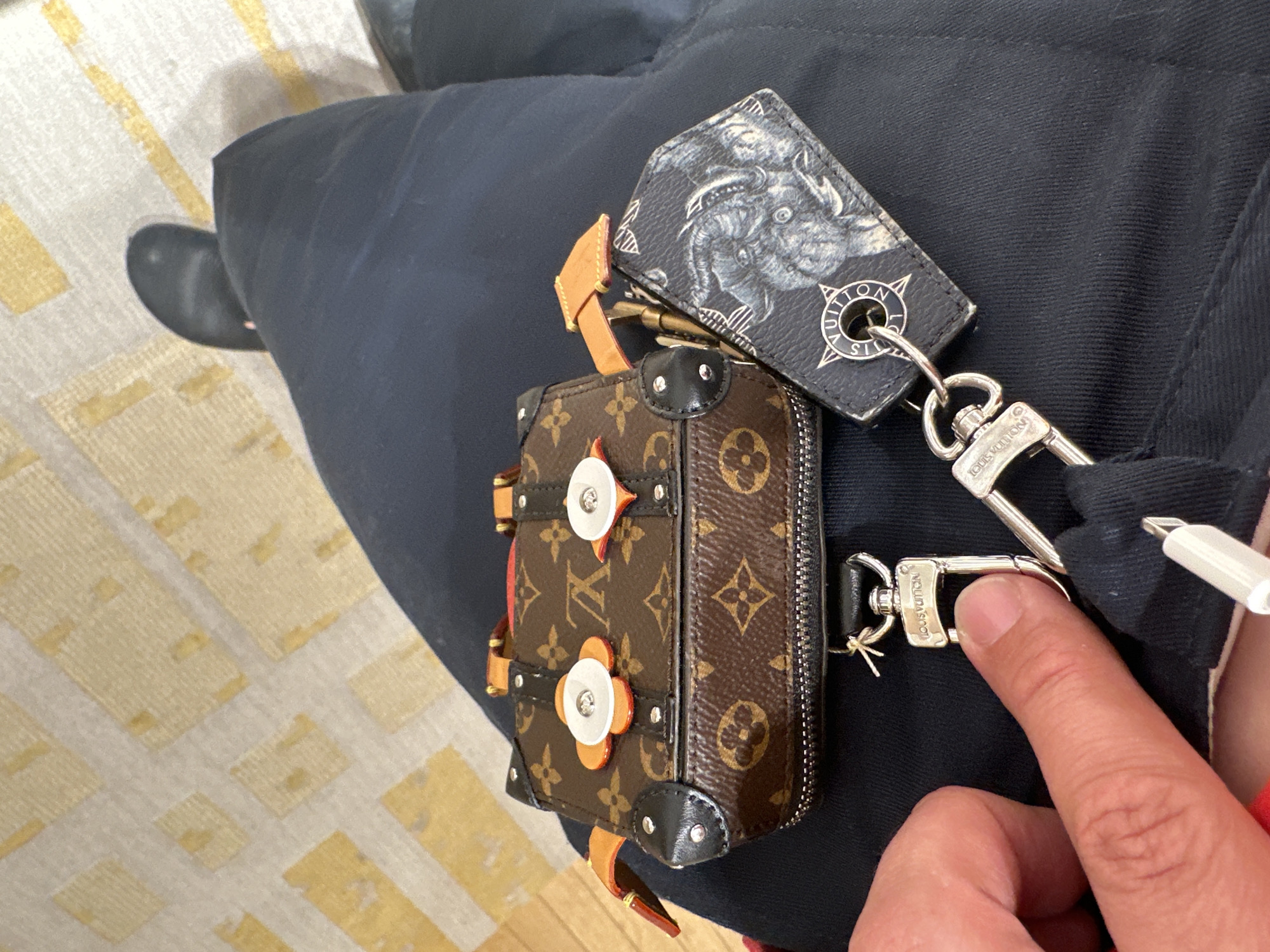 The image captures a close-up, slightly overhead view of a person's lap and hand, showcasing luxury accessories against dark attire and a patterned floor. The scene is likely indoors, with no specific time of day or weather discernible.
A prominent feature is a miniature Louis Vuitton bag, resembling a soft trunk, with its classic brown monogram canvas. It features reinforced black leather corners and distinctive orange and white flower-shaped hardware with silver studs. The bag has a zipper closure and a small, light brown leather strap.
Attached via silver-toned clasps is a rectangular black bag charm or keychain, displaying an intricate grey illustration of a creature, possibly a mythological beast or dragon. One of the circular metal rings on this charm clearly shows the "LOUIS VUITTON" brand name. A prominent silver lobster clasp, also embossed with "LOUIS VUITTON," connects the charm to the bag or an unseen item, and another silver clasp is visible near the small bag.
A person's fingers are visible in the lower right, engaging with one of the silver clasps, suggesting they are in the process of attaching or detaching an accessory. A white cable end, possibly a charging connector like a Lightning or USB-C, is also clipped or tucked nearby.
The person is wearing dark blue or black trousers, and a dark shoe is partially visible in the lower left. The floor is covered by a light-colored rug featuring a repeating pattern of yellow or beige rectangular shapes. While the location context is Madrid, Spain, there are no specific visual cues within the image to confirm this. The overall impression is one of casual display or handling of high-end fashion accessories.