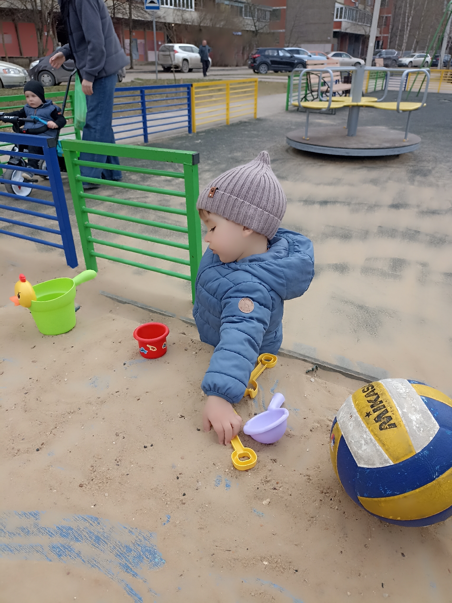 A child in a blue coat and gray hat is playing in a sandbox. The sandbox contains several plastic toys, including a green bucket, a red bucket, and yellow and purple shovels. A Mikasa volleyball is also in the sandbox. Another child is sitting in a stroller pushed by an adult. A playground with a merry-go-round is visible in the background, along with cars and buildings. The location is Serpukhov, Russia.