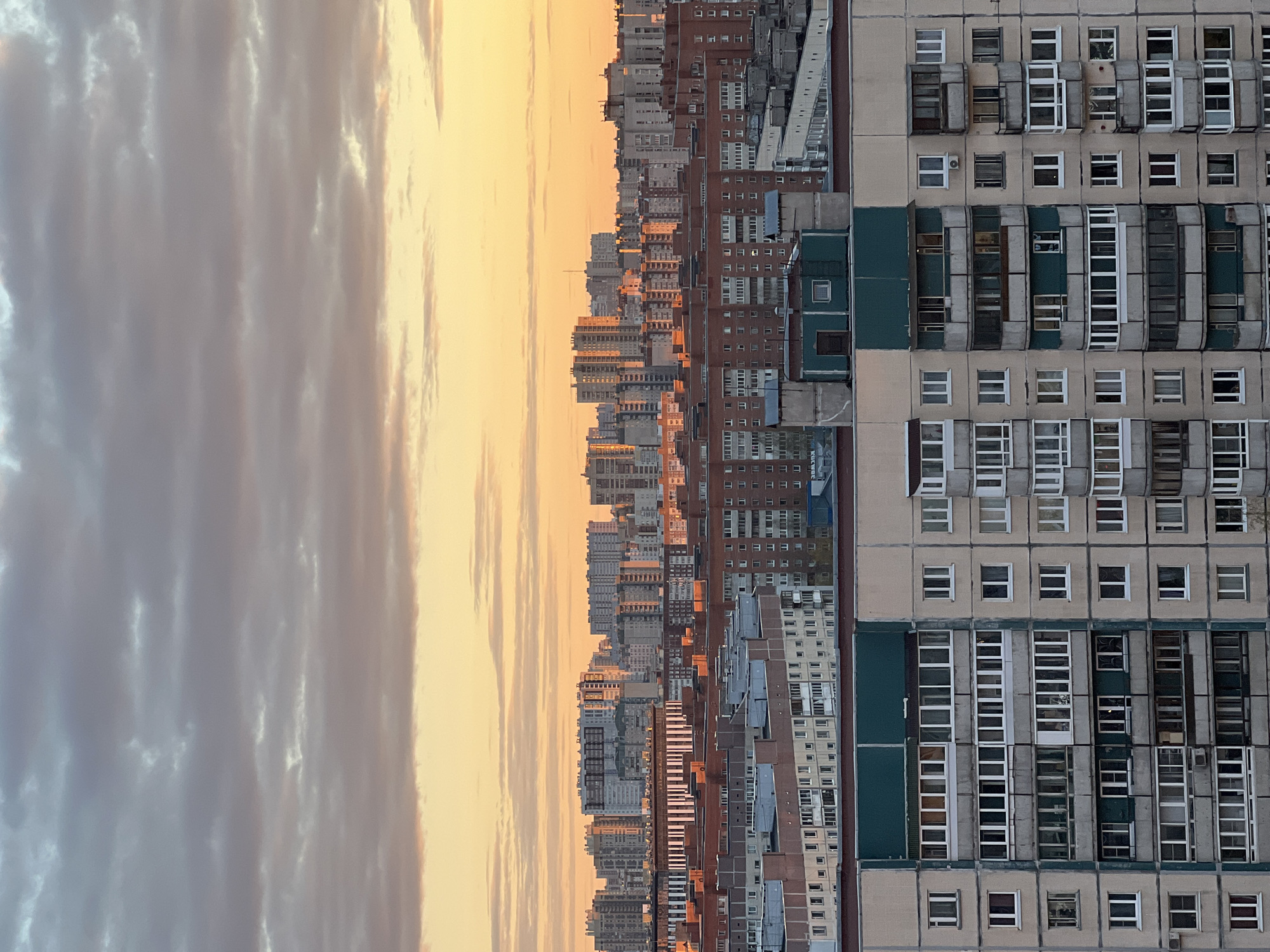The image presents an expansive urban landscape in Saint Petersburg, Russia, captured during either sunrise or sunset, from a high vantage point. The scene is dominated by a dense array of multi-story buildings under a warm, atmospheric sky.
In the foreground, occupying the right side of the frame, is a large, light-colored concrete residential building. Its facade features numerous rectangular windows and distinctive, often enclosed, balconies, some with dark green accents. Air conditioning units are visible on several windows. The building exhibits a repetitive, modular architectural style.
Beyond this structure, a vast cityscape extends into the midground and distance. A multitude of apartment blocks and high-rises form a dense horizon line, their upper sections and exposed surfaces bathed in the soft, golden light of the low sun. The urban sprawl appears continuous, characterized by various shades of grey, brown, and brick-red buildings.
The sky, occupying the left half of the image, transitions from a bright, warm golden-orange hue near the horizon, where it meets the distant city, to a softer grey-blue towards the upper portion. Long, streaky clouds, partially illuminated by the golden light, stretch across the sky, adding texture.
The overall lighting indicates either early morning or late evening, casting a serene glow over the distant city while closer elements are in partial shadow. No people are visible. On a dark green panel near the bottom right of the foreground building, there is a small, indistinct white text in what appears to be a Cyrillic or similar script, possibly reading "КУВЧ" or "KYBCH".