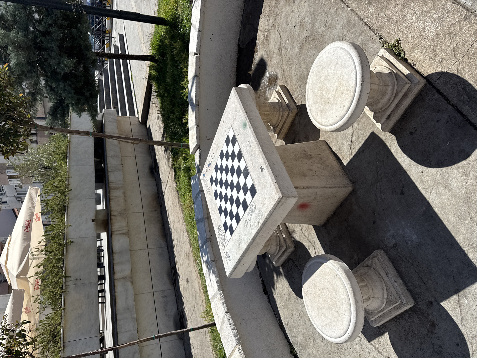 In the city of Donji Pobori, Montenegro, a public outdoor space features a stone chess table with a built-in chessboard. Two matching stone stools are positioned around the table, which is set on a concrete ground. A low, curved concrete wall surrounds the area, with grass and small trees growing behind it. In the background, there are more trees, a building, and a set of concrete stairs. The scene is bathed in sunlight, casting distinct shadows. The chess table has some graffiti markings on its surface.