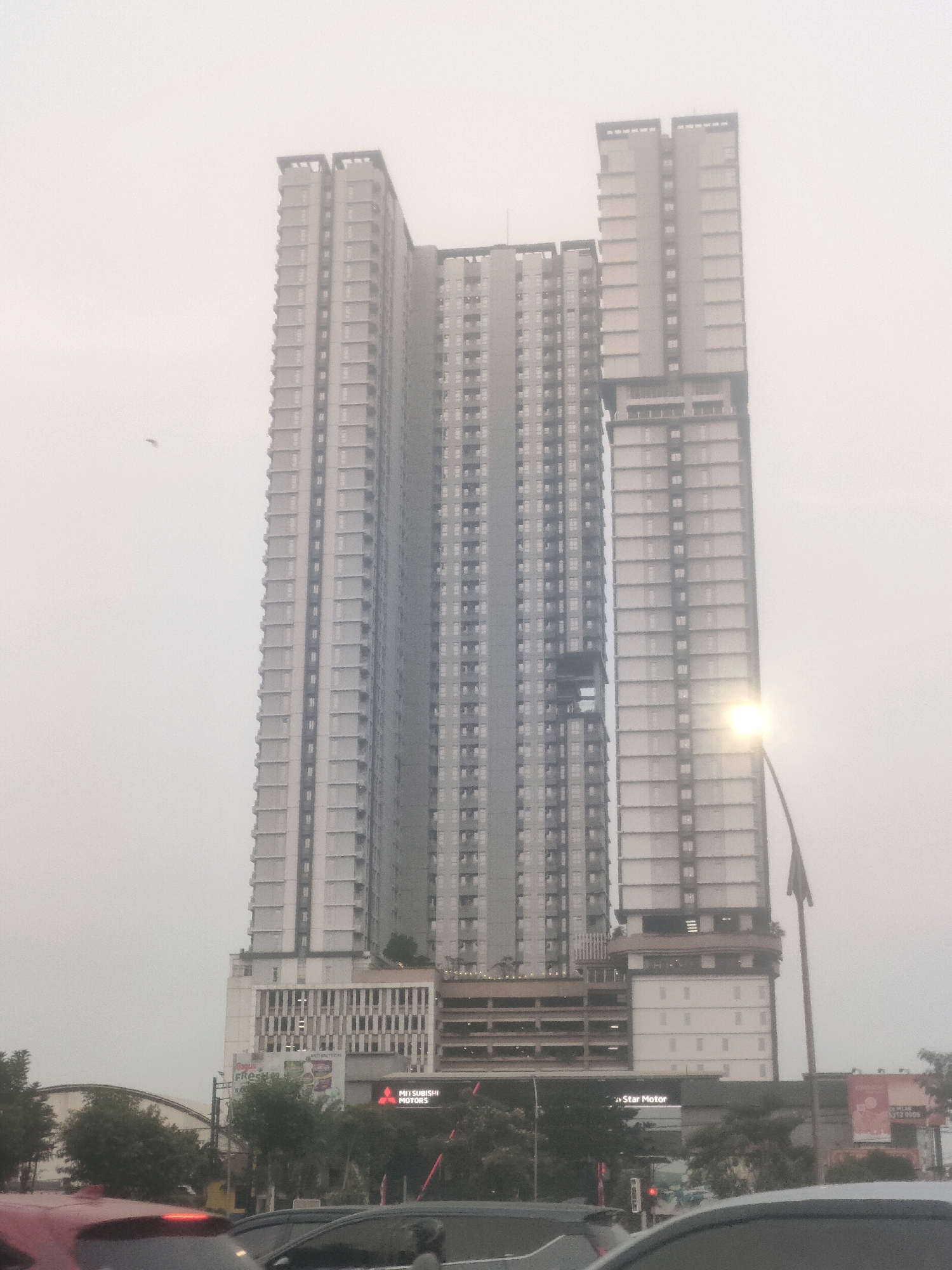 The image shows a cluster of modern, high-rise buildings in Surabaya, Indonesia, shrouded in a hazy or foggy atmosphere. The most prominent structure is a very tall, multi-sectioned skyscraper with a light-colored facade, featuring numerous windows and balconies. To its right, another tall building, slightly shorter and with a darker facade, stands. The lower sections of the buildings appear to house commercial establishments, with visible signage including "MITSUBISHI MOTORS". A street lamp is illuminated to the right of the taller building. In the foreground, the tops of cars are visible, suggesting the photograph was taken from a street level, possibly during traffic. Green trees are scattered around the base of the buildings. The overall impression is one of a dense urban environment with towering architecture, set against a muted, overcast sky.