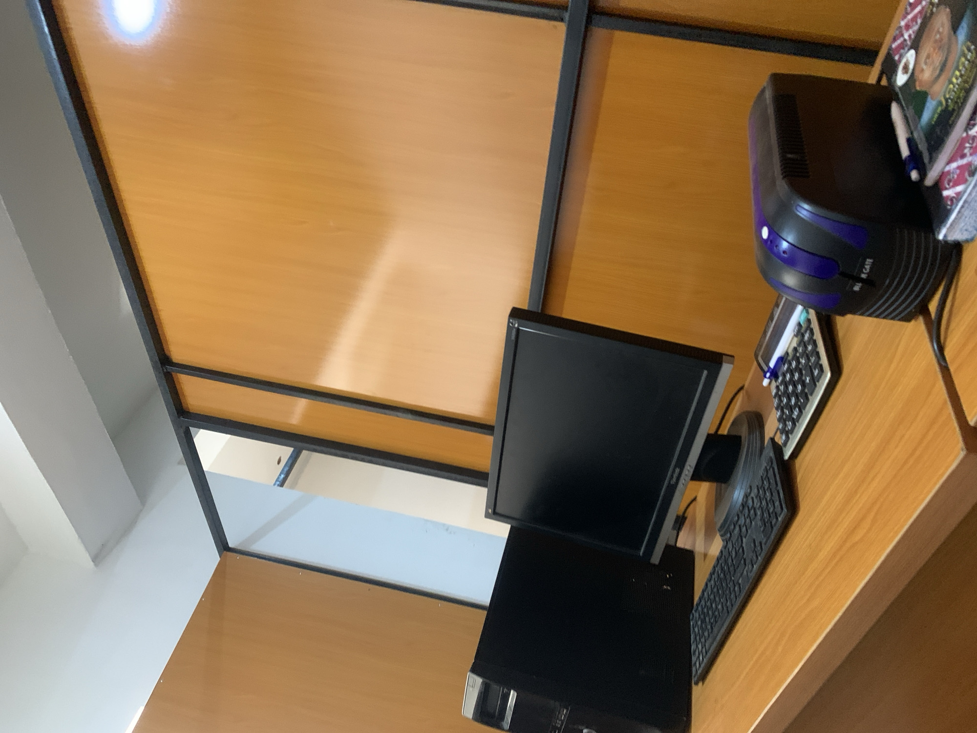 An interior scene shows a desk with a computer, monitor, keyboard, calculator, and a black and purple uninterruptible power supply (UPS). The desk is made of wood and is positioned in front of a wall with wooden panels and black metal dividers. The ceiling is white. There are no people visible in the image. The overall impression is that of a functional workspace.