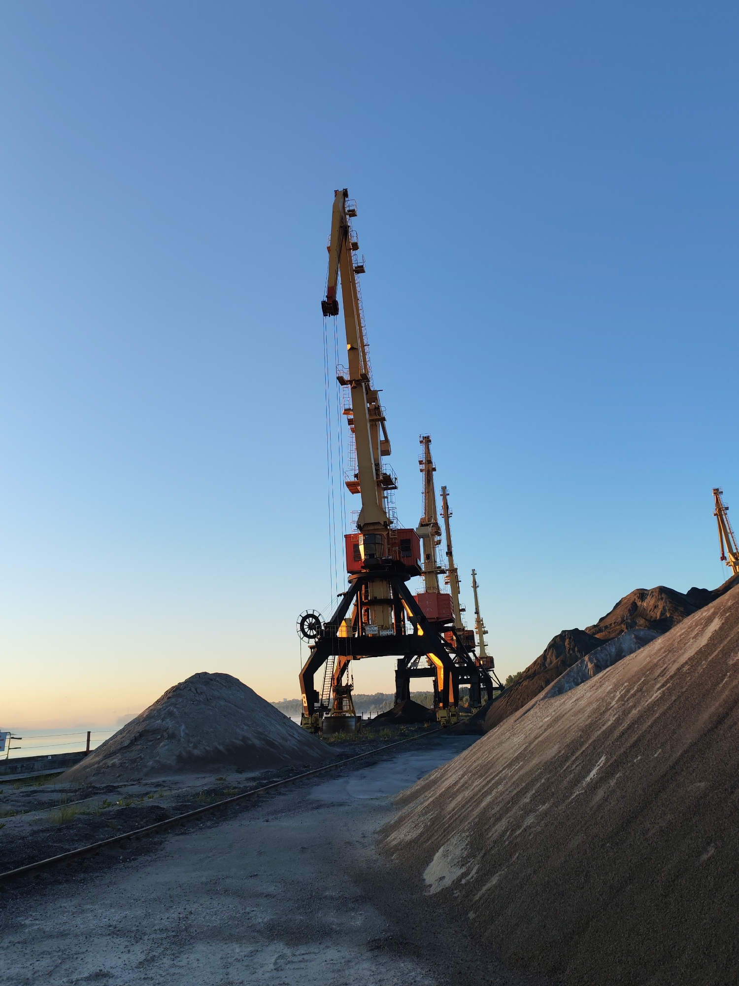 This is a low-angle, full shot of a port facility under a clear blue sky at sunrise. The image features several large, yellow and red construction cranes, their booms extended upwards. In the foreground, there are large piles of gravel and sand. Train tracks run between the piles and the cranes. In the background, there is a body of water and a hazy shoreline. The location is the port of Bol'shie Mkhi, Russia. There are no people or animals visible in the image. The overall scene is industrial and barren, with the rising sun casting long shadows and highlighting the textures of the materials.