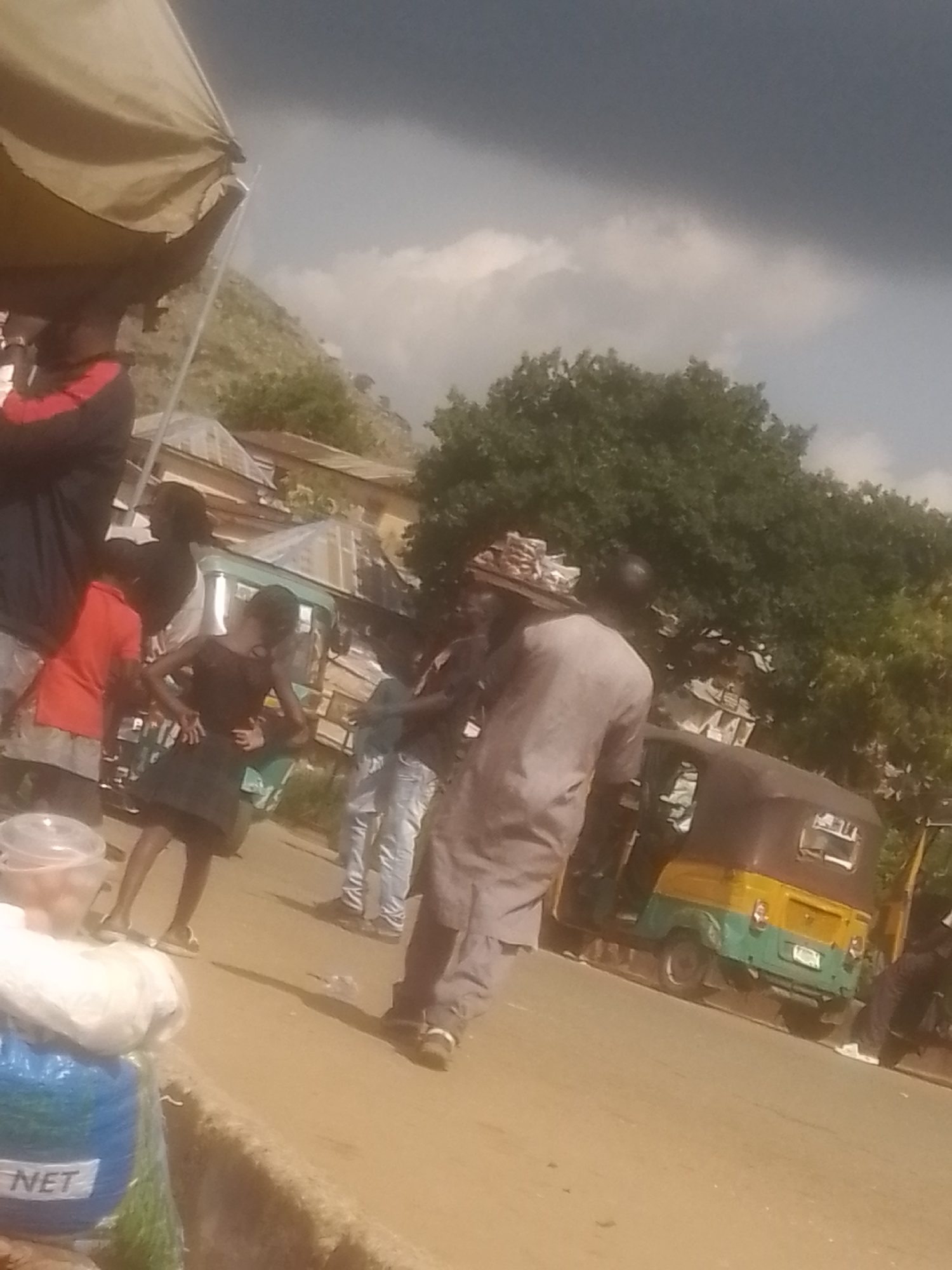 In Gwong, Nigeria, a street scene unfolds under a dramatic, cloudy sky. A large, tan awning casts a shadow over part of the foreground, where a person in a dark jacket with red accents stands. Further back, a group of individuals are gathered. A woman in a black dress dances, her hands on her hips, while other people, including a man in a light gray traditional outfit, observe. A green and yellow auto-rickshaw, commonly known as a "Keke," is parked nearby. In the foreground, a plastic container filled with items sits beside a blue plastic bag with the word "NET" visible on it. The backdrop features buildings with corrugated roofs and lush green trees, suggesting a semi-urban or rural setting.