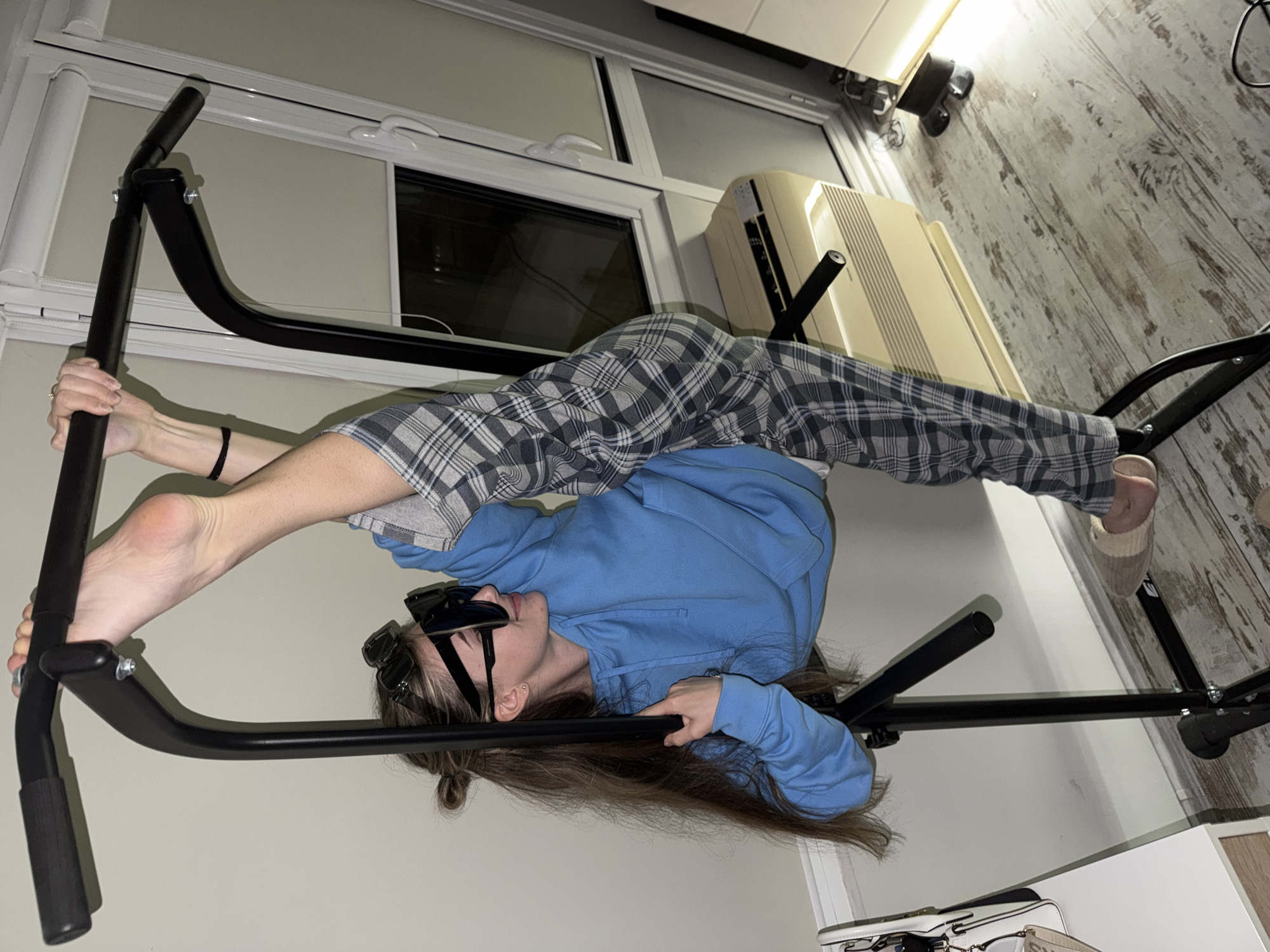 A woman wearing sunglasses, a blue hooded sweatshirt, and plaid pants is doing a split while positioned upside down on a black pull-up bar. She holds the bar with both hands and one foot, while her other foot is pointed upwards. The pull-up bar is located in a room with a window, an air conditioning unit, and light-colored walls. The floor is wood-patterned. The image doesn't show enough to determine the exact location within the city of Plovdiv, Bulgaria.