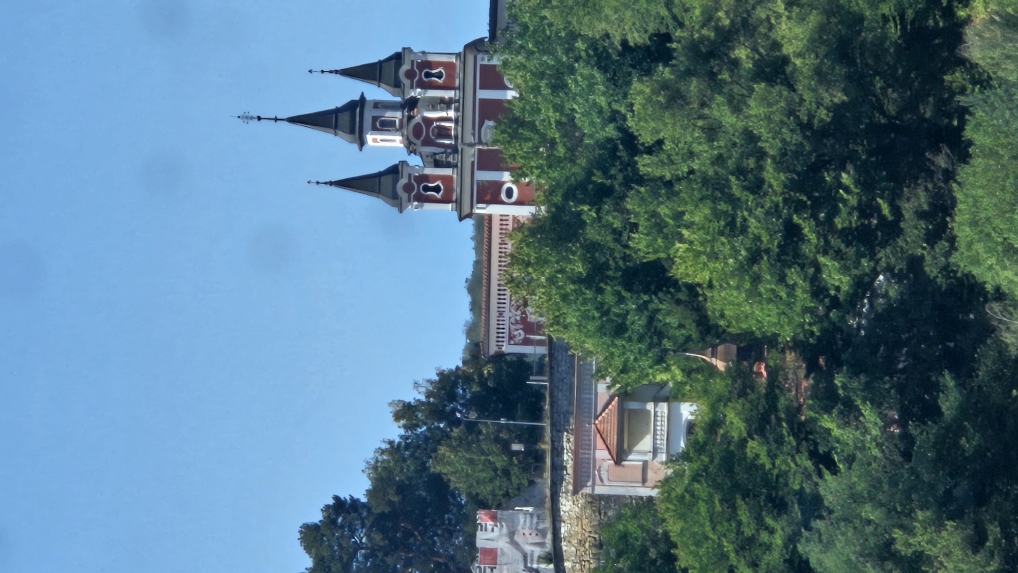 A picturesque view of the Franciscan Church and Monastery, a prominent landmark in Prešov, Slovakia, is presented. The image captures the church's distinctive red and white architecture, featuring multiple spires topped with crosses, standing against a clear blue sky. The church is nestled amidst lush green trees, with a stone wall and a smaller building with an orange roof visible in the foreground, adding to the scene's charm. The overall impression is one of serene beauty, showcasing the historic architecture and natural landscape of the city.