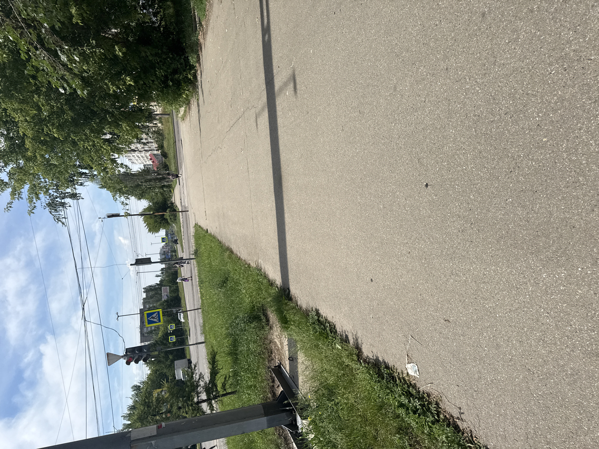 The image depicts a street scene in Angarsk, Russia, on a bright, partly cloudy day. A paved sidewalk runs diagonally across the foreground, bordered by a grassy verge on the left. In the distance, a traffic light with a red signal is visible, along with pedestrian crossing signs. Several overhead wires crisscross the sky. A line of trees and buildings can be seen along the street. Two individuals are walking in the distance, and a white truck is parked to the left. The overall impression is of a typical urban environment.