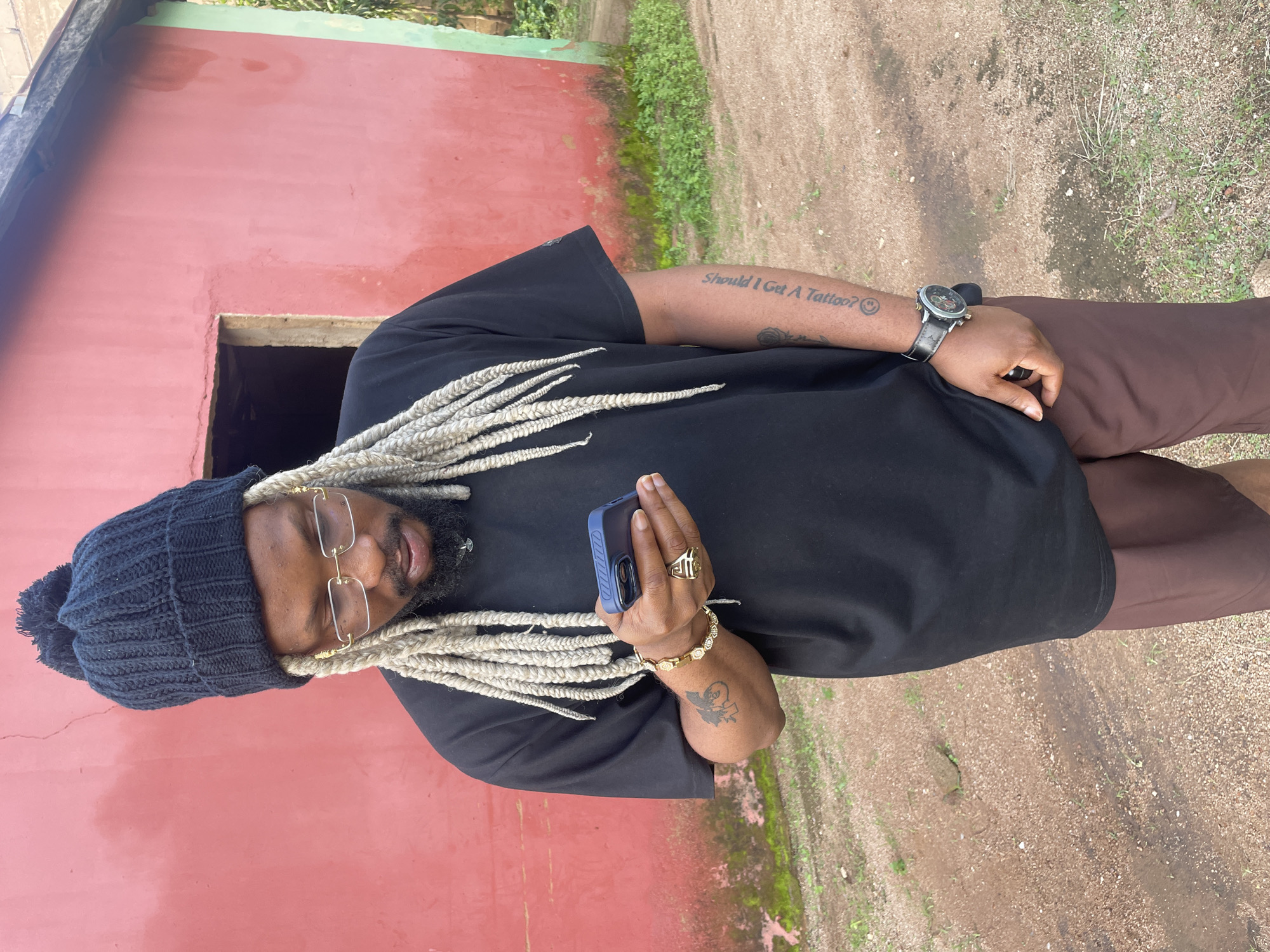 A man with light-colored braided hair, wearing glasses, a black beanie, and a black t-shirt, stands outside a red building. He is holding a dark blue phone in his right hand, and he is wearing a gold ring and a gold bracelet. His left arm has a tattoo of a rose and text that reads "Should I Get A Tattoo?". He also has a watch on his left wrist. The background shows a dirt path and some greenery. The image is taken during the daytime, and the weather appears to be fair.