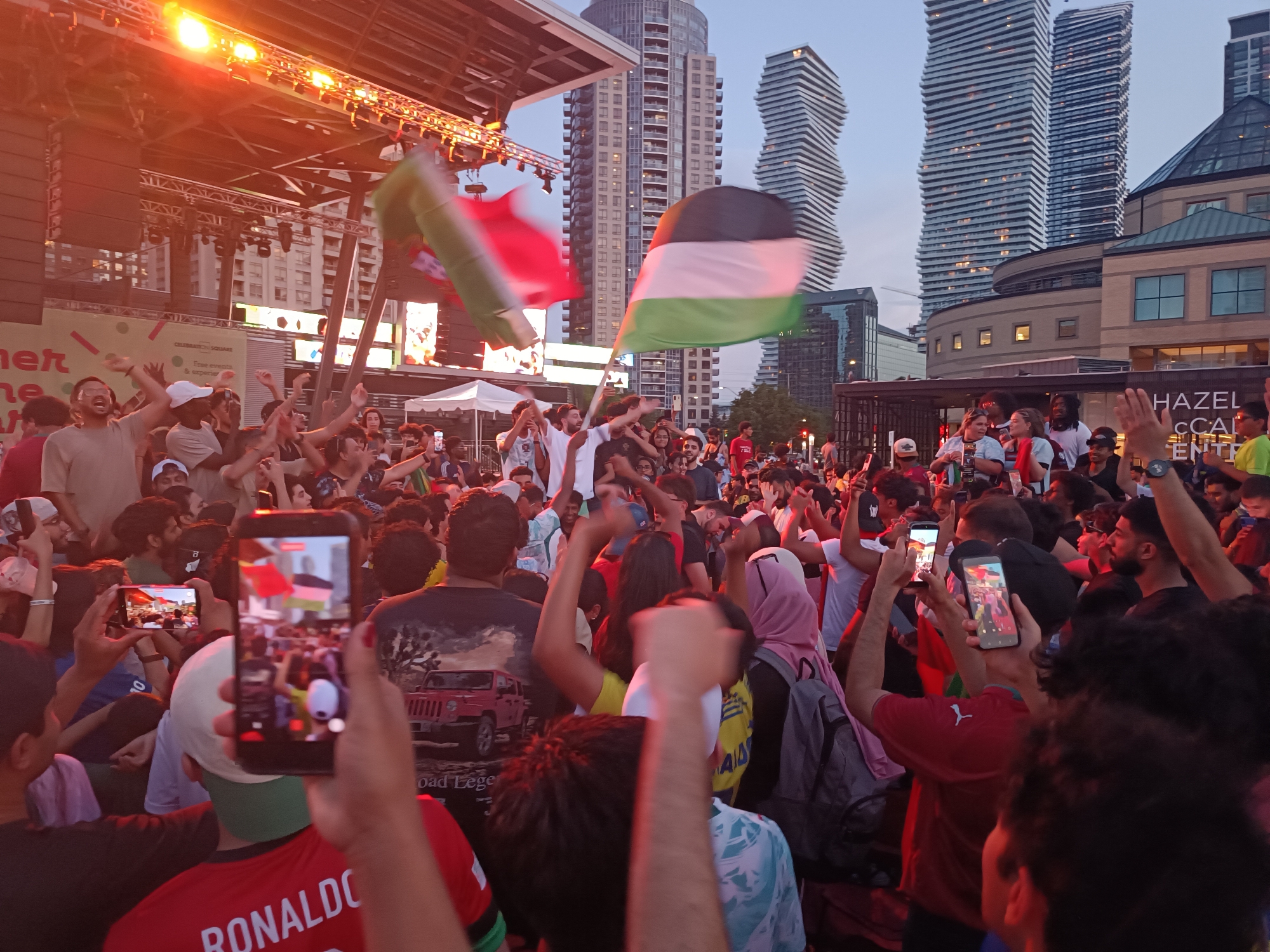 This is a medium shot, eye-level, outdoor photograph taken at Celebration Square in Mississauga, Canada, capturing a lively crowd gathered for what appears to be an event. The sky is a dusky blue, indicating either early evening or late afternoon. In the background, distinctive modern skyscrapers, including the distinctive twisting towers of Mississauga City Centre, rise against the sky.
In the foreground and mid-ground, a dense crowd of people are visible, many with their arms raised in the air, suggesting excitement and engagement with an event taking place on a stage or platform to the left. The stage is illuminated by bright yellow stage lights, and speaker systems are mounted above. Two flags are prominent in the frame: a red, white, and green flag (possibly a Palestinian flag) is held aloft on the left, and a larger flag with horizontal stripes of black, white, green, and red (also likely a Palestinian flag) is waving on the right.
Several people are holding up smartphones, recording or photographing the event. One person in the foreground is holding a red t-shirt with "RONALDO" printed on the back in white lettering. Another individual's t-shirt displays a graphic of a red Jeep. A woman in the mid-ground is wearing a pink headscarf. The overall atmosphere is one of energetic celebration, with a diverse group of people participating. The lighting is warm, cast by the stage lights and the setting sun, creating a vibrant and dynamic scene.