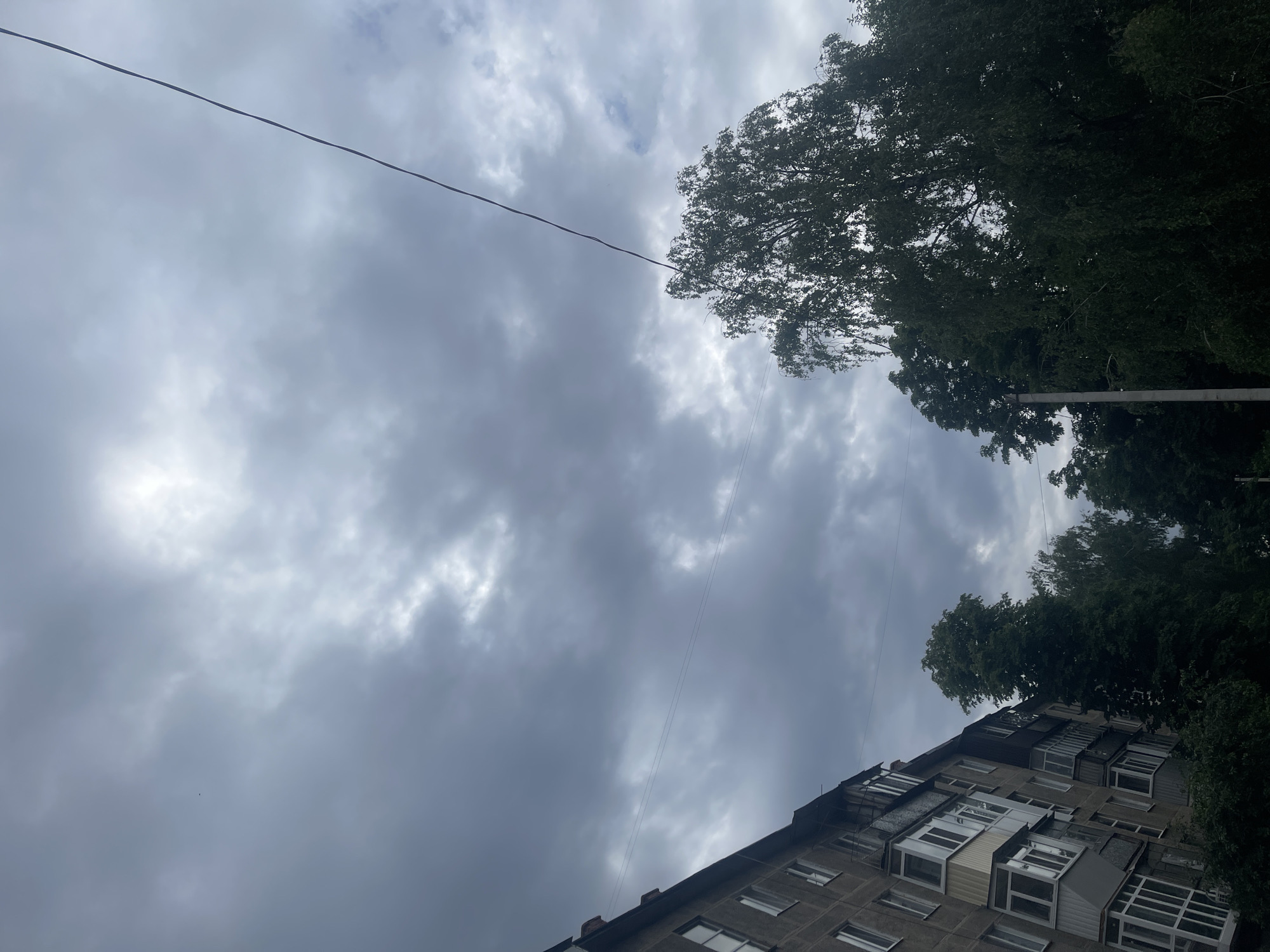 The image captures an upward-angled view of an urban environment in Poltava, Ukraine, on an overcast day. The majority of the frame is dominated by a cloudy, grey sky, with some brighter areas indicating diffused sunlight breaking through the dense cloud cover.
A prominent dark utility wire stretches diagonally across the upper portion of the sky. To the lower right, a multi-story apartment building with a distinctive architectural style, typical of post-Soviet residential buildings, is partially visible. The building's facade features numerous windows and a variety of enclosed and open balconies or loggias, some appearing to be updated or customized.
On the right side of the image, lush green deciduous trees stand tall, their dense foliage contrasting with the grey sky and partially obscuring the building. There are no people visible, nor any specific activities taking place, presenting a static, everyday scene. The weather appears cloudy and possibly humid. No visible text is present in the image.