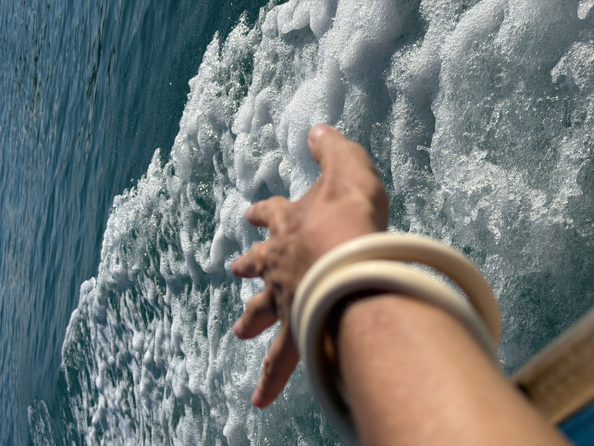 This media file captures a dynamic perspective of a hand reaching into frothy, turquoise water, likely from a moving vessel. The scene is dominated by the churning wake of a boat, creating a texture of white foam and sparkling water droplets. The hand, adorned with two light-colored bangles on the wrist, is blurred due to motion, suggesting the speed of the water or the camera. The fingers are outstretched, seemingly about to touch or interact with the water.
The setting appears to be a bright, sunny day, with the sunlight glinting off the water and foam, creating a sense of energy and movement. The clear, deep turquoise of the undisturbed water to the left contrasts with the white, effervescent wake, highlighting the motion. This perspective offers a sense of immersion and the exhilaration of being on the water. The location context of Baan Thong Krut, Thailand, evokes a tropical marine environment. No text is visible in the media file. The overall mood is one of active engagement with the surrounding natural elements.