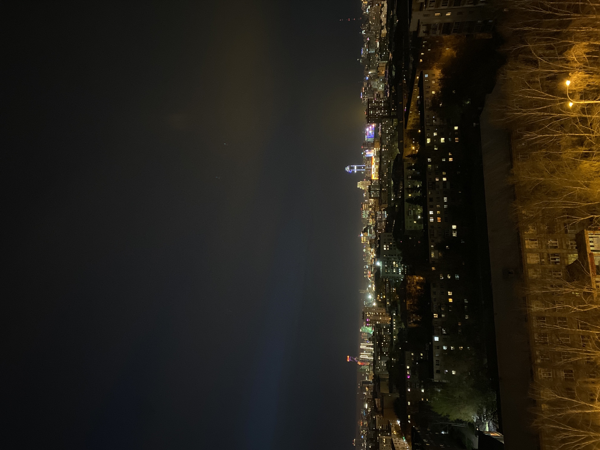 This is a nocturnal wide-angle view of the city of Yekaterinburg, Russia, captured from a high vantage point, likely a tall building or elevated terrain. The image depicts an expansive urban landscape stretching into the distance under a dark, hazy night sky.
In the immediate foreground on the right, several bare, leafless tree branches are prominently visible, strongly illuminated by a bright, warm yellow streetlight. The branches suggest a season like late autumn, winter, or early spring. Adjacent to these trees, the dark facade of a building is partially visible, hinting at the structure from which the photo might have been taken or a neighboring one.
The midground showcases a dense collection of buildings, both residential and commercial, adorned with numerous artificial lights. Many windows glow with warm yellow and white light, indicating activity within. A notable detail is a modern, taller building in the mid-distance, featuring distinctive, vibrant blue, purple, and pink architectural lighting, making it a focal point against the predominantly warm-toned city lights. Other buildings exhibit various forms of facade lighting and illuminated signs.
Towards the background, the city extends far into the horizon, with countless lights forming a glowing expanse that gradually fades into the atmospheric haze and darkness. The sky above is a deep charcoal grey to black, showing no stars, likely due to light pollution from the city. No people or specific activities are discernible, and there is no visible text. The overall scene conveys a sense of quiet urban grandeur at night.