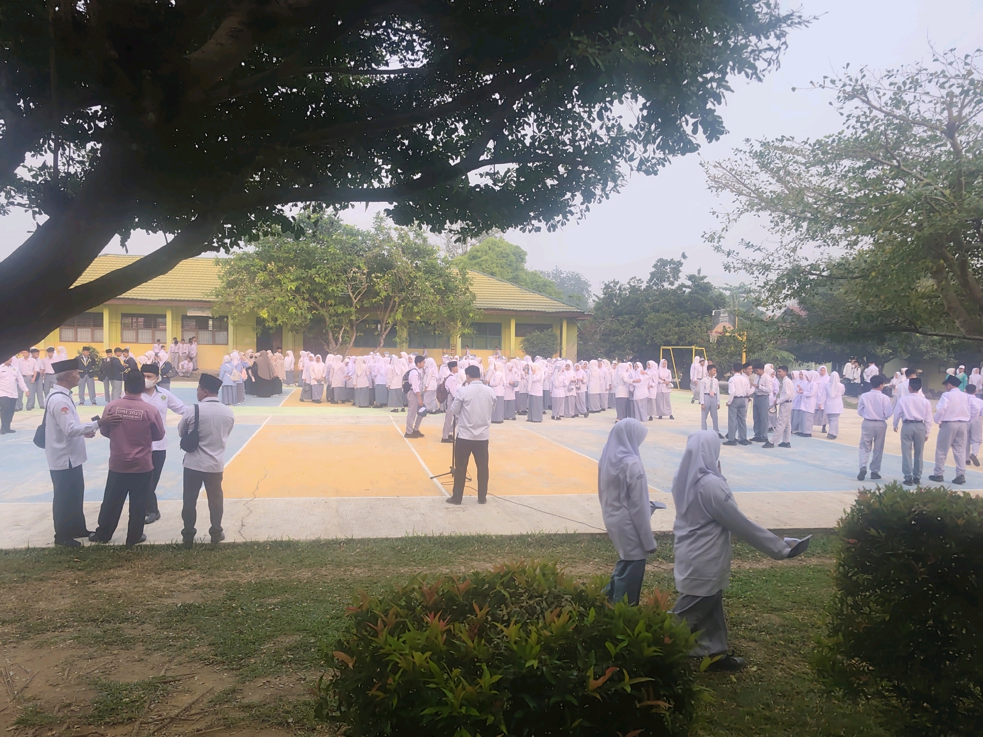 In Pematang Reba, Indonesia, a group of students, predominantly in white uniforms, are gathered on an outdoor court, possibly for a school event. The court is marked with colored lines, suggesting it may be a sports facility. A building with a yellow roof and green walls is visible in the background. Several adults are also present, some appearing to be teachers or organizers, interacting with the students. The scene is set under a large tree, with lush green foliage in the foreground and surrounding the court. The atmosphere appears to be orderly and community-oriented, with many individuals standing in formation or engaged in conversation.
