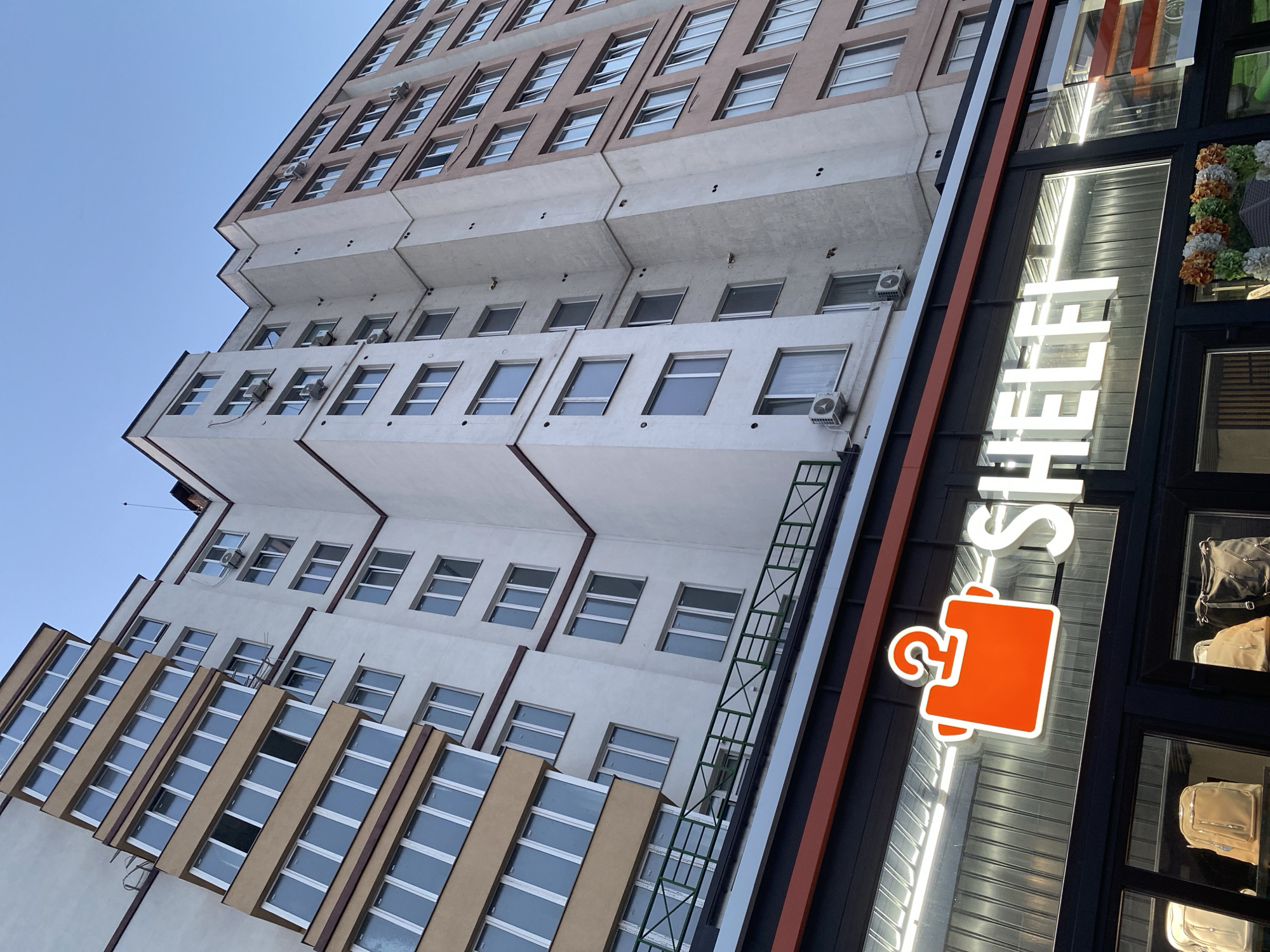 The image shows a multi-story residential building with a store on the ground floor. The building has a light-colored facade with multiple windows and balconies. On the ground floor, there is a store with a bright orange logo featuring a hanger with a question mark above a stylized shirt. The store name "SHELFI" is written in large white letters. The windows of the store display several bags. The scene appears to be an exterior shot of a commercial and residential area. Based on the provided information, this location is in the city of Vinnytsia, Ukraine.