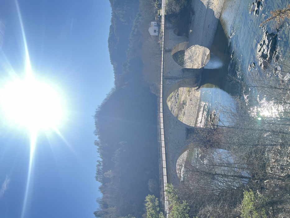 The image displays a multi-arch stone bridge spanning a river under a clear, intensely bright blue sky, with the sun prominently centered in the upper frame, generating significant lens flare. This scene is located in Stoianovo, Bulgaria. The bridge, constructed of light-colored masonry, features a wooden handrail along its top and extends across a river with visible shallow areas, rocky banks, and sandbars. The river water is clear, reflecting sunlight and sky. On the left side, evergreen foliage and bare deciduous trees are visible in the foreground, with densely forested hills rising in the background. On the right side of the bridge, a small, white, single-story building stands on the riverbank, accompanied by additional green structures further along the bank. No individuals, vehicles, or animals are present.