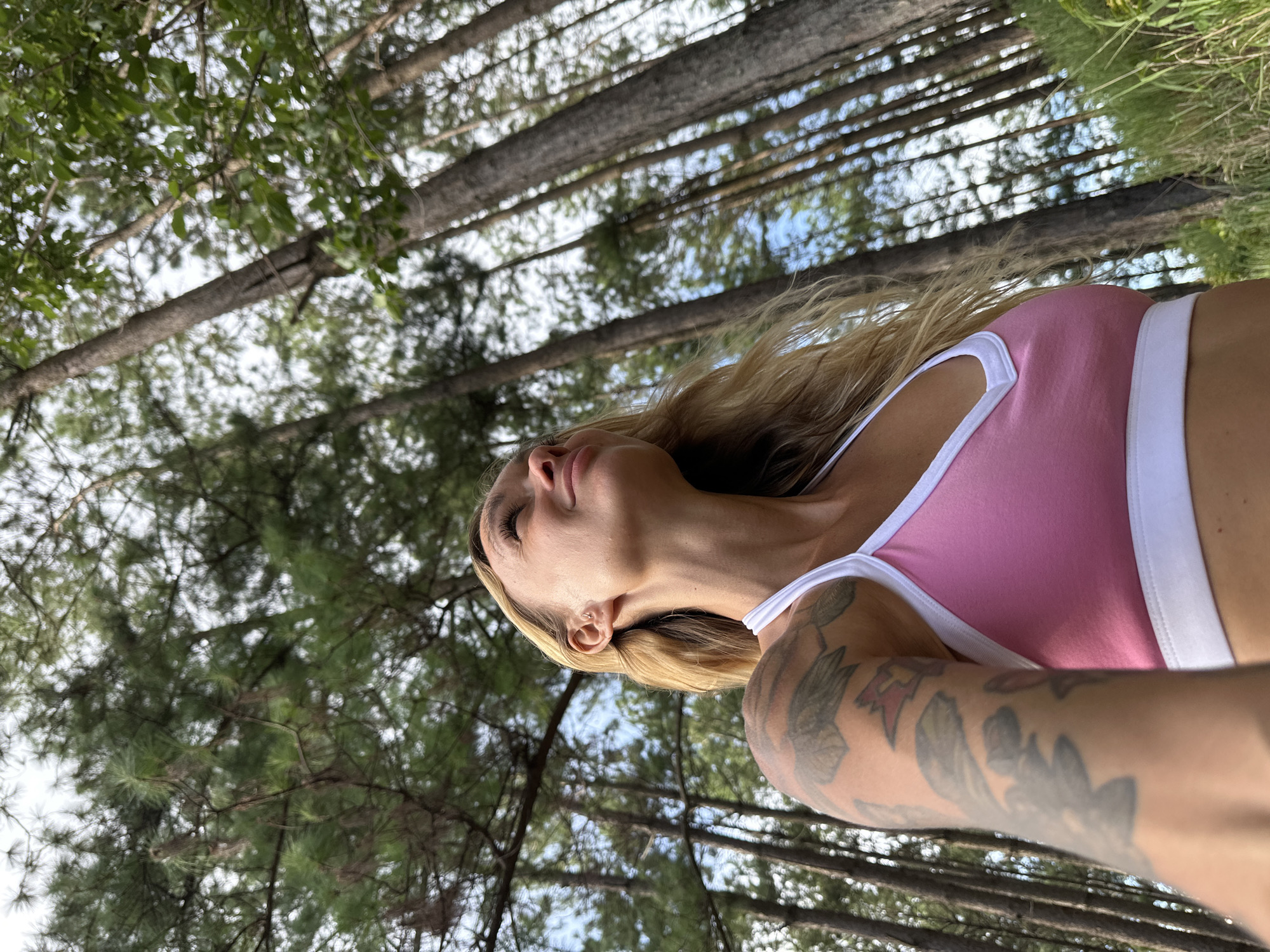 A woman with blonde hair and tattoos on her arm is shown from the chest up, looking upwards with her eyes closed. She is wearing a pink sports bra with white trim. The background is out of focus and consists of trees and greenery, suggesting an outdoor setting. The lighting suggests it is daytime. The image is oriented vertically with the top of the frame showing the sky through the trees.
