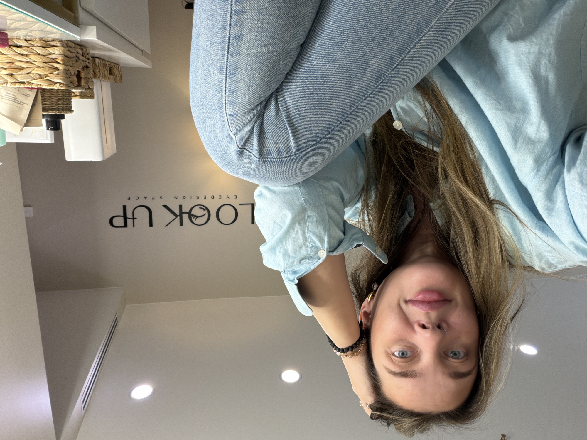 A young woman with light skin, blue eyes, and blonde hair is looking directly at the camera. She is wearing a light blue denim shirt and blue jeans. She has a nose ring and is wearing bracelets and earrings. The background includes a light beige wall with the words "LOOK UP EYE DESIGN SPACE" written on it. There is a white shelf with a woven basket and various items on it. There are recessed ceiling lights. There is no indication that the location is in Moscow, Russia.
