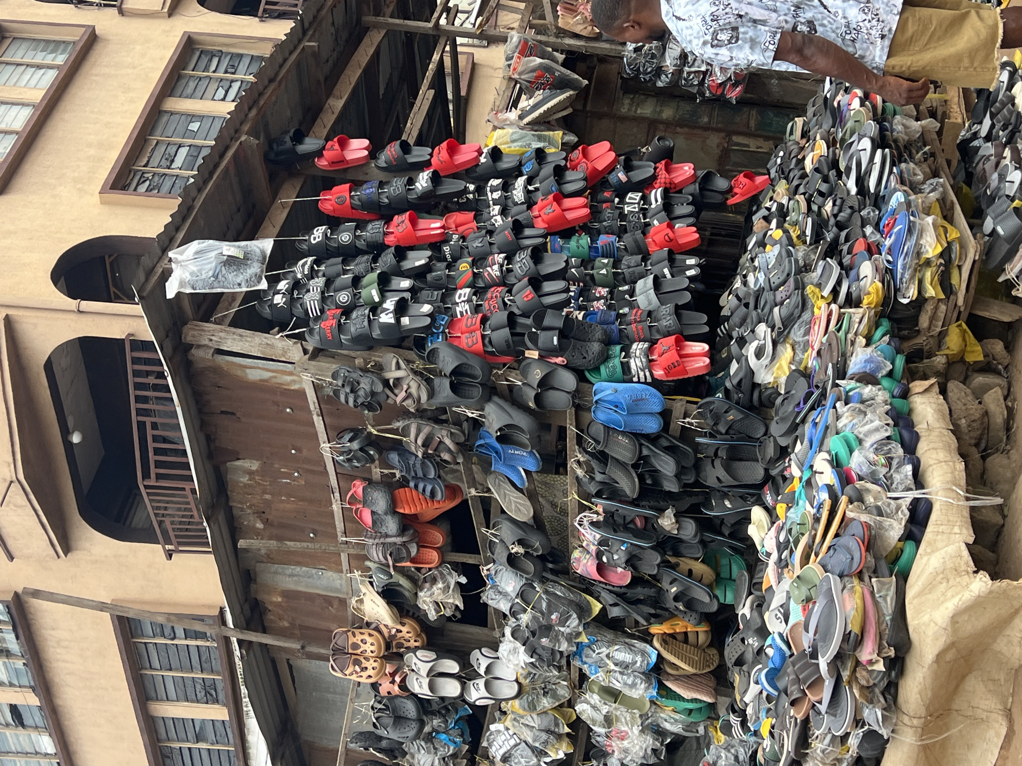 The image depicts a vibrant outdoor market stall in Nkpor, Nigeria, overflowing with a diverse array of footwear. The stall itself appears to be constructed from corrugated metal sheeting and wooden planks, suggesting a temporary or informal setup. Suspended from makeshift racks are hundreds of flip-flops, sandals, and slides in various colors and styles, including popular brands and generic designs. Some footwear is packaged in plastic, while others are displayed individually.
The stall is situated in front of a multi-story building with a beige facade, featuring numerous windows with broken panes and dark metal frames. The architecture suggests a blend of residential and commercial use, common in urban Nigerian settings. In the foreground, a man wearing a patterned shirt and khaki shorts is partially visible, his back turned to the camera, as he appears to be browsing or interacting with the stall owner. The ground is covered with a rough canvas material, and piles of footwear are neatly stacked on wooden pallets. The overall impression is one of a bustling marketplace, characteristic of Nkpor's commercial activity.