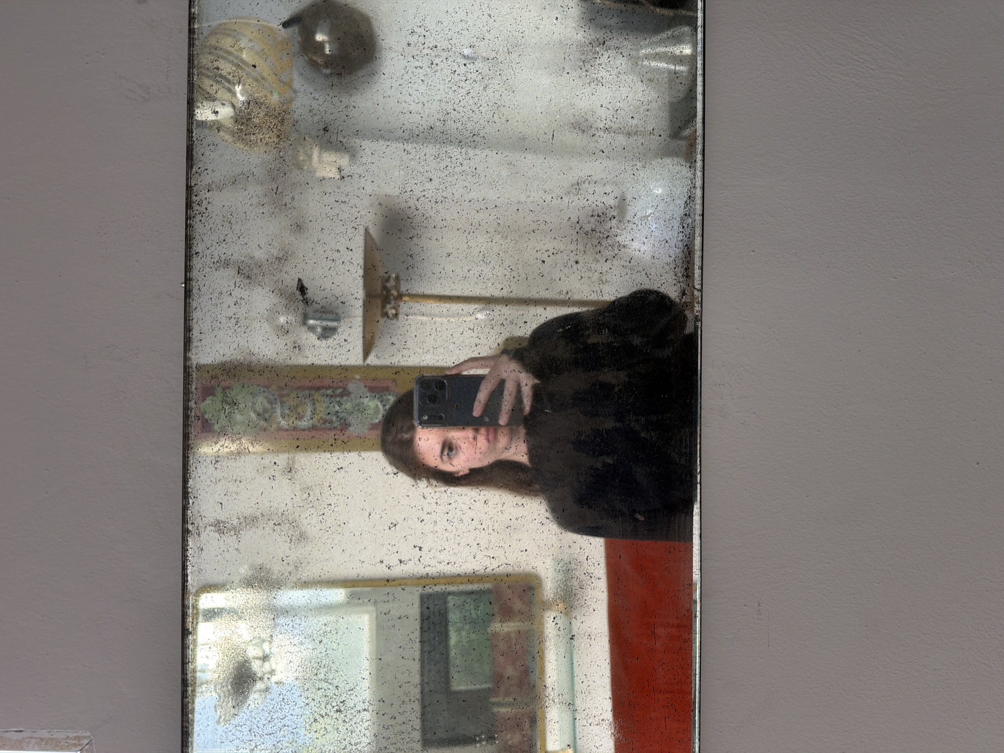 An indoor, vertical image captures a selfie reflected in a heavily distressed and aged mirror. The mirror occupies the left portion of the frame, with a neutral, light gray wall on the right.
The primary subject is a person, likely female, whose reflection is seen holding a smartphone to take the picture. She is oriented horizontally within the vertical frame, with her head positioned towards the left edge and her body extending to the right. She has dark hair and is wearing a dark top or sweater. Her expression is neutral as she focuses on the phone. The smartphone appears to be a modern model with multiple cameras.
The mirror's surface is a notable feature, showing significant signs of age with widespread speckling, dark spots, and areas where the silvering appears to have deteriorated, creating a textured, almost antique effect. This aged surface partially obscures the reflections.
Reflected in the mirror are glimpses of the indoor environment:
* In the upper section, vague shapes of decorative objects or fixtures are visible against a lighter background.
* Mid-frame, a golden-colored, wall-mounted fixture with a flat, rectangular component is reflected.
* In the lower part of the mirror, another reflection shows what appears to be part of an interior space, possibly another mirror or a window, with a white frame.
* A vibrant red surface or object is reflected on the right side, partially behind the person's reflection.
The activity is a mirror selfie being taken indoors. No specific time of day or weather is discernible. The setting suggests an interior space, possibly in an older building consistent with location contexts such as Buenos Aires, Argentina. No visible text is present in the image.