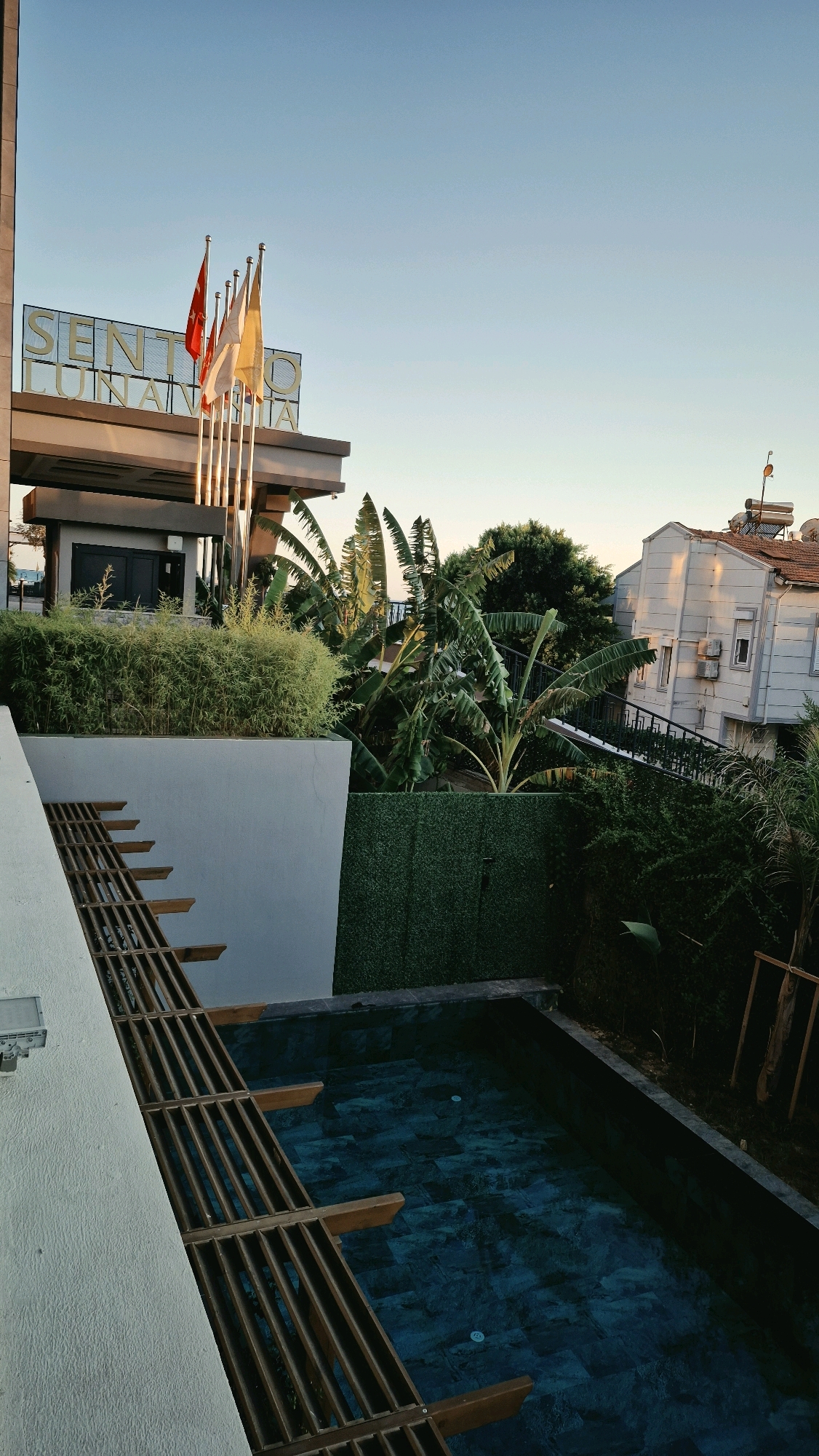 The image depicts the exterior of a hotel or resort, likely in Serik, Turkey, with a clear blue sky. In the foreground, a white textured wall with a wooden slatted structure runs diagonally. To the right, a dark tiled swimming pool is visible, with what appears to be a dark green artificial hedge bordering it. Behind the hedge, lush green vegetation, including large-leafed banana plants, dominates the mid-ground. To the far right, a multi-story building with white siding, a terracotta-tiled roof, and solar panels is partially visible. In the background, there are more trees and buildings. On the left side of the image, the entrance to the establishment is marked by a sign with large golden letters spelling out "SENTO LUNARIA," with several flags flying above it. The scene suggests a warm climate and a well-maintained outdoor area, possibly during the late afternoon or early morning due to the lighting. There are no people visible in the image.