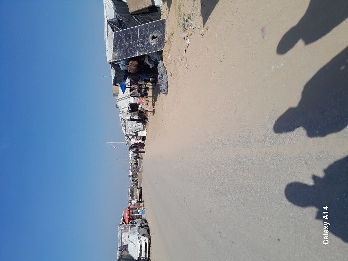 The image depicts a wide-angle view of a bustling makeshift settlement alongside a road in Khan Yunis, Palestinian Territory, under a clear blue sky during daylight hours. A two-lane asphalt road, partially covered with dust and sand, runs from the foreground into the distance, appearing less defined in the background.
On the left side of the road, a series of crude shelters constructed from tarps, fabric, and other scavenged materials line the roadside. Several vehicles are parked here, including a white car completely covered with a light-colored sheet or tarp, and another sedan immediately behind it. Further down, individuals are seen walking between structures, and a small market stall with a red and white striped umbrella is visible.
On the right side of the road, a dense row of similar makeshift shelters, fashioned from black, white, and blue tarps, fabric, and boards, extends into the distance. Prominently in the mid-ground, a large solar panel, dark in color, stands upright and leans against a shelter; it exhibits a circular impact hole near its center. Various items, including bags and clothing, hang from the exteriors of these shelters, suggesting dwellings or small commercial activities. Multiple individuals are present along this side of the road, moving and standing near the shelters.
In the immediate foreground, distinct shadows of at least three individuals, presumably including the photographer, are cast onto the road surface. The bright sunlight indicates midday or early afternoon conditions. The overall scene suggests an area densely populated by displaced individuals, with basic infrastructure and visible daily life activity. The "Galaxy A14" watermark is visible in the lower left corner.