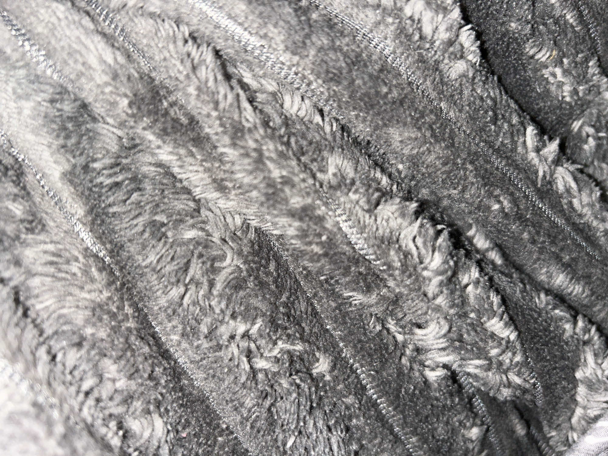 A close-up, high-detail view of a textured textile material is depicted, located in Gauting, Germany. The fabric features parallel, diagonal ridges of a light grey, subtly reflective material, approximately 1-2 cm in width, separated by areas of darker grey, dense, shaggy pile. The pile exhibits an uneven, matted texture with visible fibers and shadows indicating depth, suggesting a soft, plush surface. The overall color palette is monochromatic in shades of grey. No persons, specific objects beyond the fabric, or discernable actions or interactions are present. The immediate environment is solely defined by the textile's surface.
