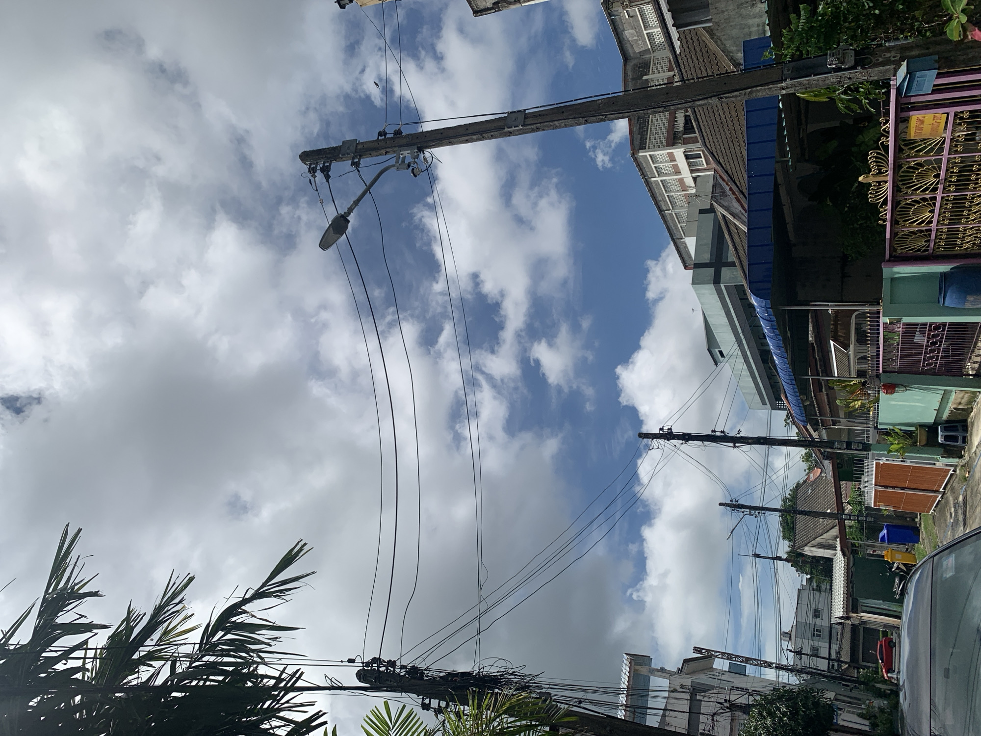 The image depicts a residential street in an urban environment under a cloudy sky with patches of blue. A cluster of utility poles with numerous overhead wires dominates the center and left side of the frame. Palm fronds extend into the upper left corner, casting shadows. Buildings of various sizes and architectural styles are visible on both sides of the street, including a multi-story building with white gridded windows in the background. In the foreground, the hood of a dark-colored car is visible. To the right, a row of houses features covered patios, fences, and a bright yellow sign with Thai script. Two blue and yellow trash bins are present near a pair of orange gates. The overall impression is of a dense urban setting with a strong overhead network of electrical and communication lines.
