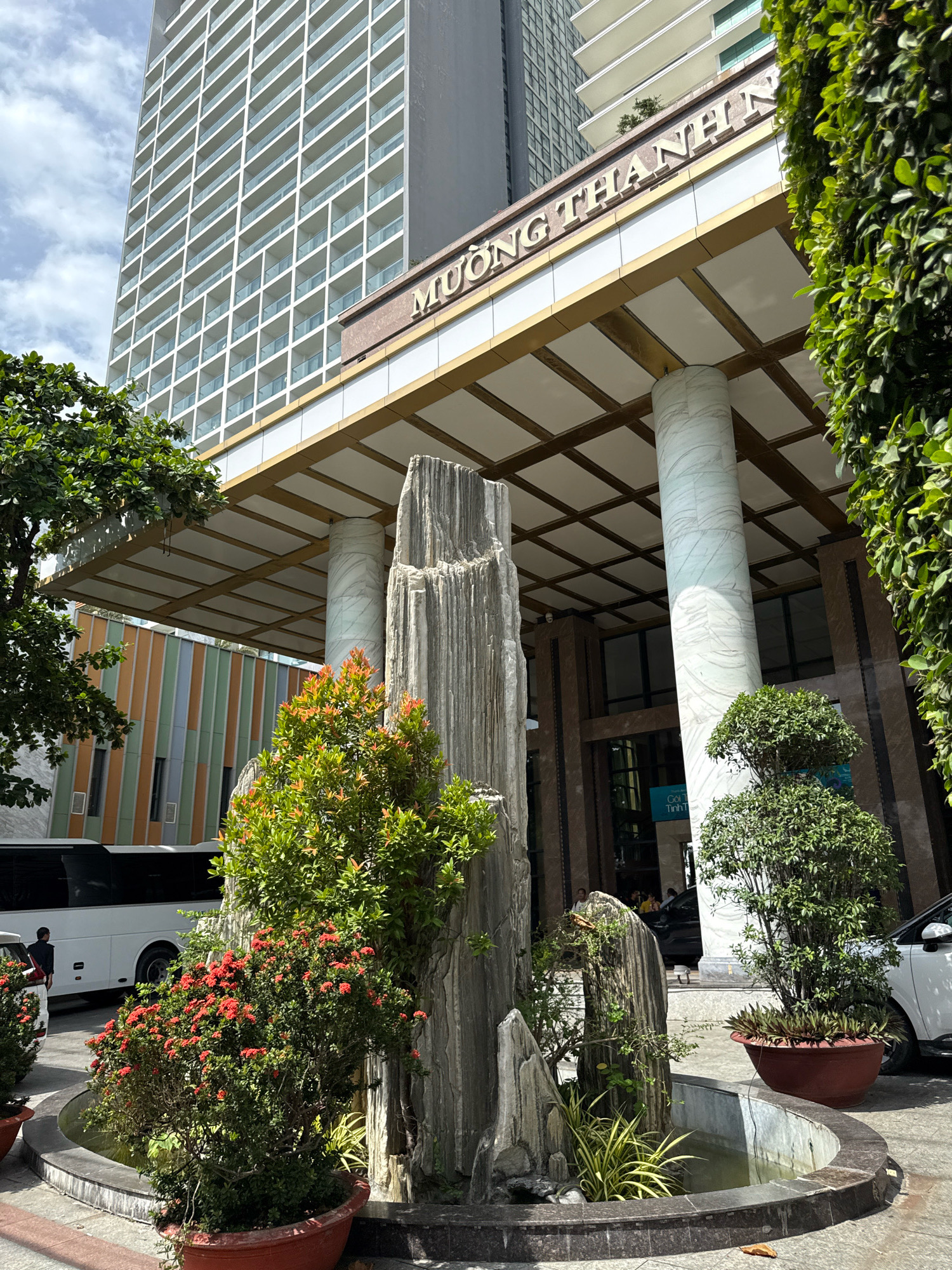 The image depicts the entrance to the Muong Thanh Nha Trang Hotel in Nha Trang, Vietnam. A modern, multi-story hotel with a white grid facade dominates the background. The hotel's name, "MUONG THANH NHN," is visible in gold lettering above the entrance. The entrance features large marble columns and a covered walkway with a white tiled ceiling. In the foreground, a decorative rock formation with lush green plants and red flowers is situated in a circular water feature. To the left of the rock formation, a white bus is parked, and a person in a dark uniform is visible near it. To the right, a white car is parked. The scene is illuminated by daylight, suggesting it was taken during the day. The weather appears to be partly cloudy.