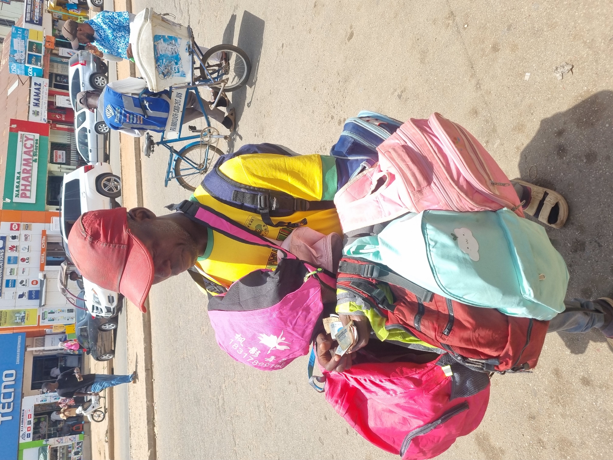 The image shows a man on a street in Jos, Nigeria, weighed down by a large pile of colorful backpacks and other bags. He is wearing a red baseball cap and a yellow shirt. He is standing in front of shops with signs for "TECNO," "NASARA PHARMACY & Stores," and "HAMAZ." In his hands, he holds a stack of 1000 Naira notes.
In the background, another man is seen riding a bicycle with a white box attached to the front. Several cars are parked along the street. The setting appears to be a bustling commercial area.