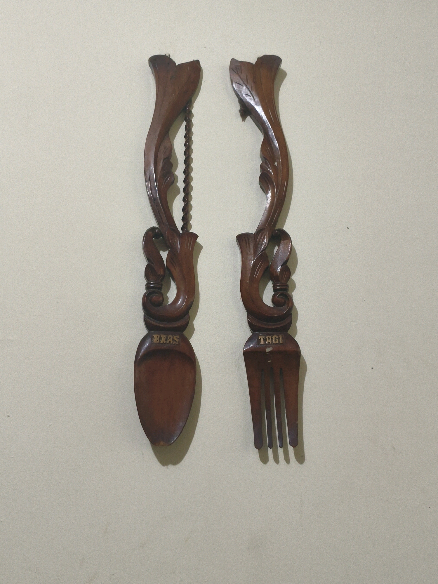 A close-up, eye-level, vertical shot displays a pair of ornate, carved wooden utensils, likely decorative, hanging against a plain, textured, light-colored wall. The utensil on the left resembles a spoon, while the one on the right is a fork. Both are crafted from dark wood and feature intricate carvings along their handles, including swirling leaf motifs and beaded detailing on the spoon's chain. The spoon is inscribed with the word "BIAS" in gold lettering on its bowl, and the fork is inscribed with "TAGI" in gold lettering on its tines. The lighting is soft, casting subtle shadows from the objects onto the wall. The overall impression is one of artistic craftsmanship displayed as wall decor. This image was taken in Bogor, Indonesia.