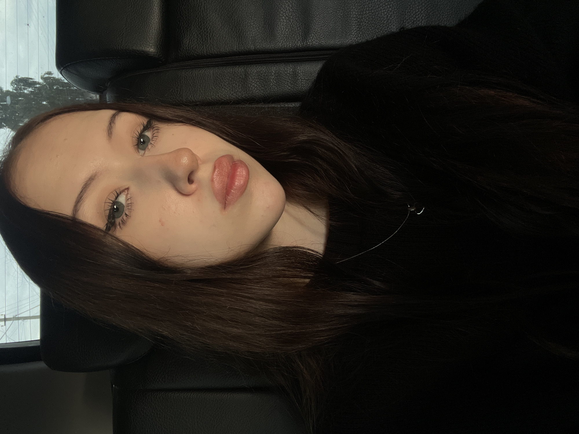 A young woman with long, dark brown hair and green eyes is shown in a close-up selfie. She is wearing a black sweater and a silver necklace with a small pendant. Her lips are slightly pursed. She appears to be sitting in the backseat of a vehicle, with black leather upholstery visible behind her. The background shows a window with a glimpse of trees and power lines, suggesting an outdoor setting in a semi-urban or rural area. The lighting is dim, with the primary light source coming from the front, illuminating her face. There is no indication of her specific location within Gavrilovka, Russia, beyond being in a vehicle there.