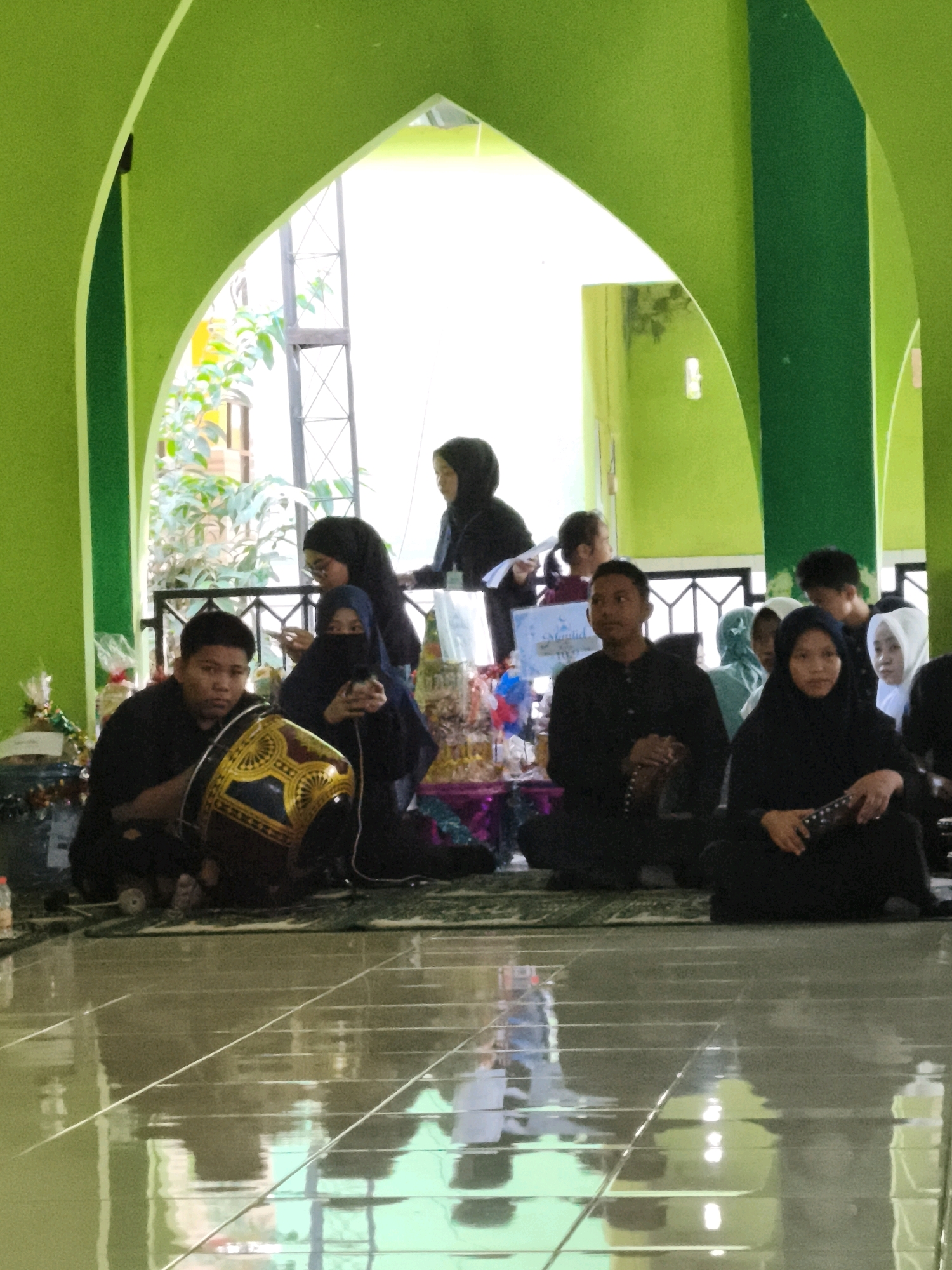 The image depicts a gathering of people, predominantly women in hijabs and men in black clothing, inside a building with vibrant green arched architecture, suggesting a religious or community event, possibly a mosque or community hall, in Makassar, Indonesia. In the foreground, a young man sits with a traditional drum adorned with yellow and black patterns. To his right, several women are seated, some wearing face veils. Behind them, more individuals are visible, including children. The floor appears to be tiled and highly reflective, mirroring the green architecture and the figures present. The background shows windows or open archways revealing greenery and daylight. Several gift-wrapped items are placed to the left and right of the main group. The atmosphere suggests a solemn or celebratory occasion.