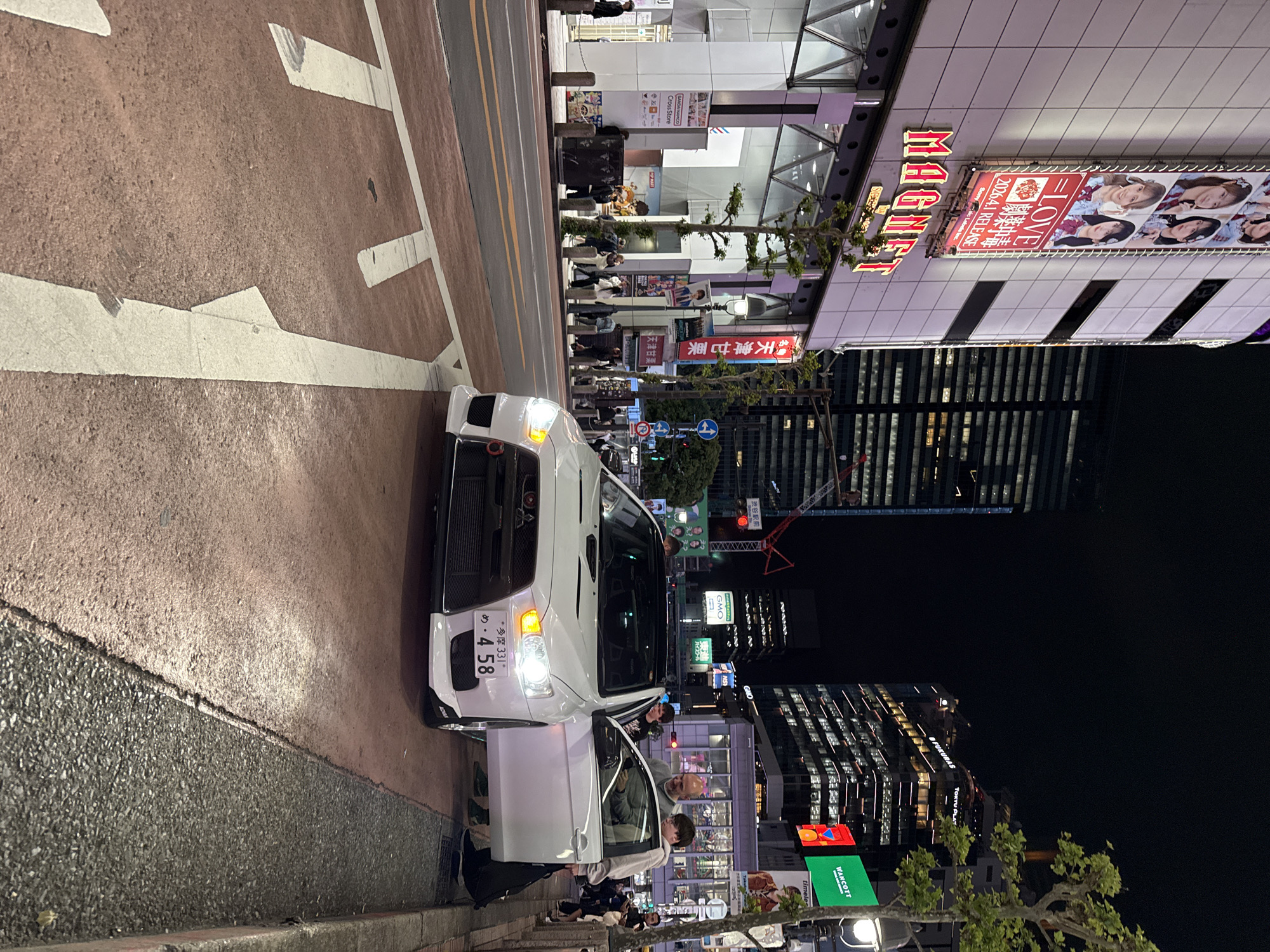The image depicts a nighttime scene in Shibuya, Japan, centered around a white car parked on the side of a road. People are seen around the car and on the sidewalk. The car's headlights are on, and a door is open. In the background are tall buildings, illuminated signs, and streetlights. A sign with the word "MAGNET" and another with "LOVE 2026.4.1 RELEASE" are visible on the buildings. There are advertisements and signs for businesses such as BANDAI NAMCO Cross Store. Street markings and pedestrian walkways are also present.