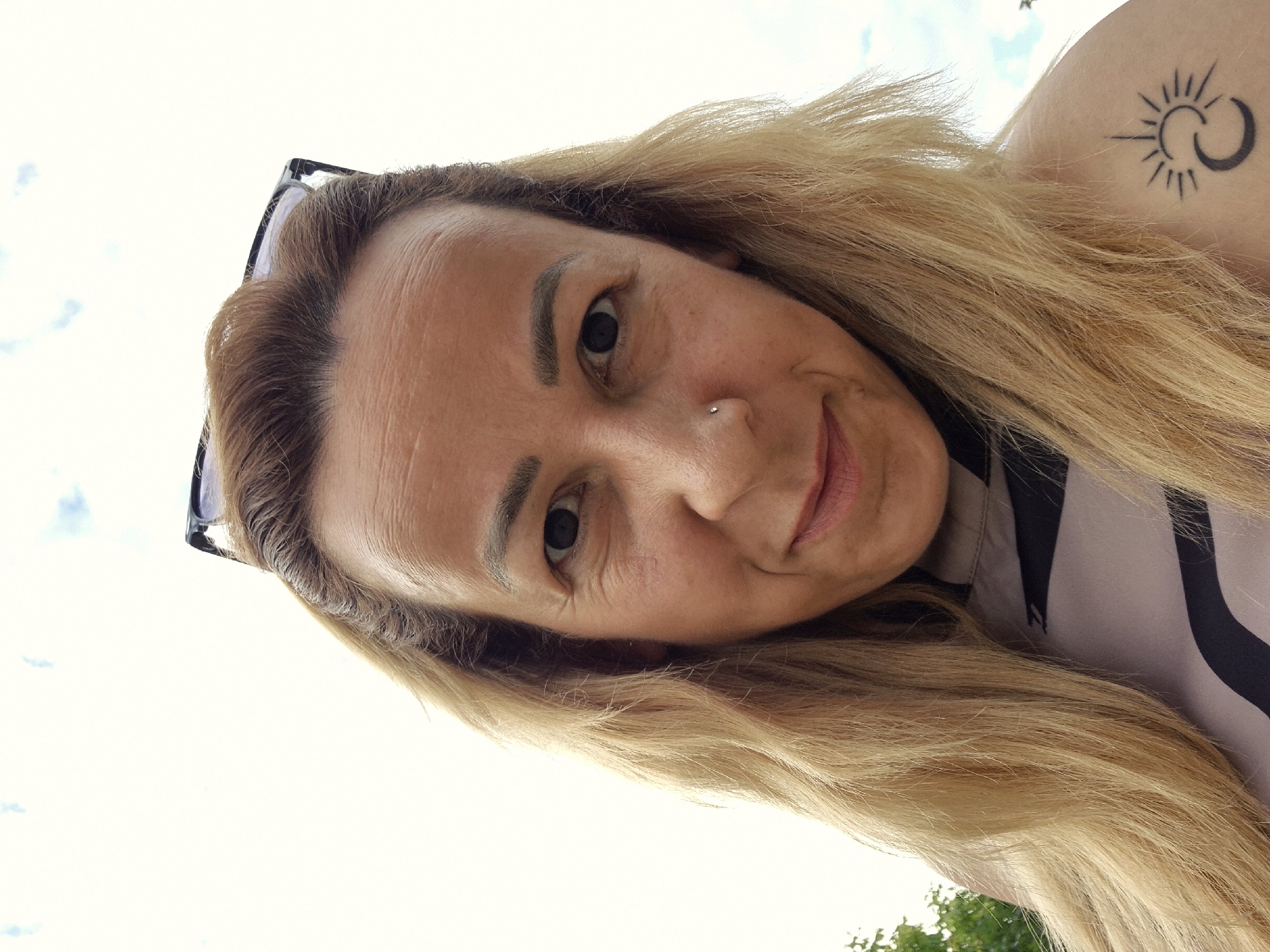 A close-up, low-angle selfie depicts a woman outdoors under a bright, largely clear sky with faint white clouds. The woman, appearing to be middle-aged with light skin and long, wavy blonde hair, occupies the majority of the frame. Her dark-rimmed glasses are pushed up onto the top of her head. Visible facial features include pronounced horizontal lines on her forehead, wrinkles around her dark eyes, and a small silver stud piercing on her left nostril. She has well-defined dark eyebrows and a slight, closed-mouth smile. Her light-colored top with dark striped patterns is visible on her torso. A distinct black tattoo is visible on her left shoulder, illustrating a stylized sun with rays emanating from it, intertwined with a crescent moon. Green foliage is partially visible on the far left and right edges of the background. The specific location in the city of Unknown, Unknown cannot be determined from the image.