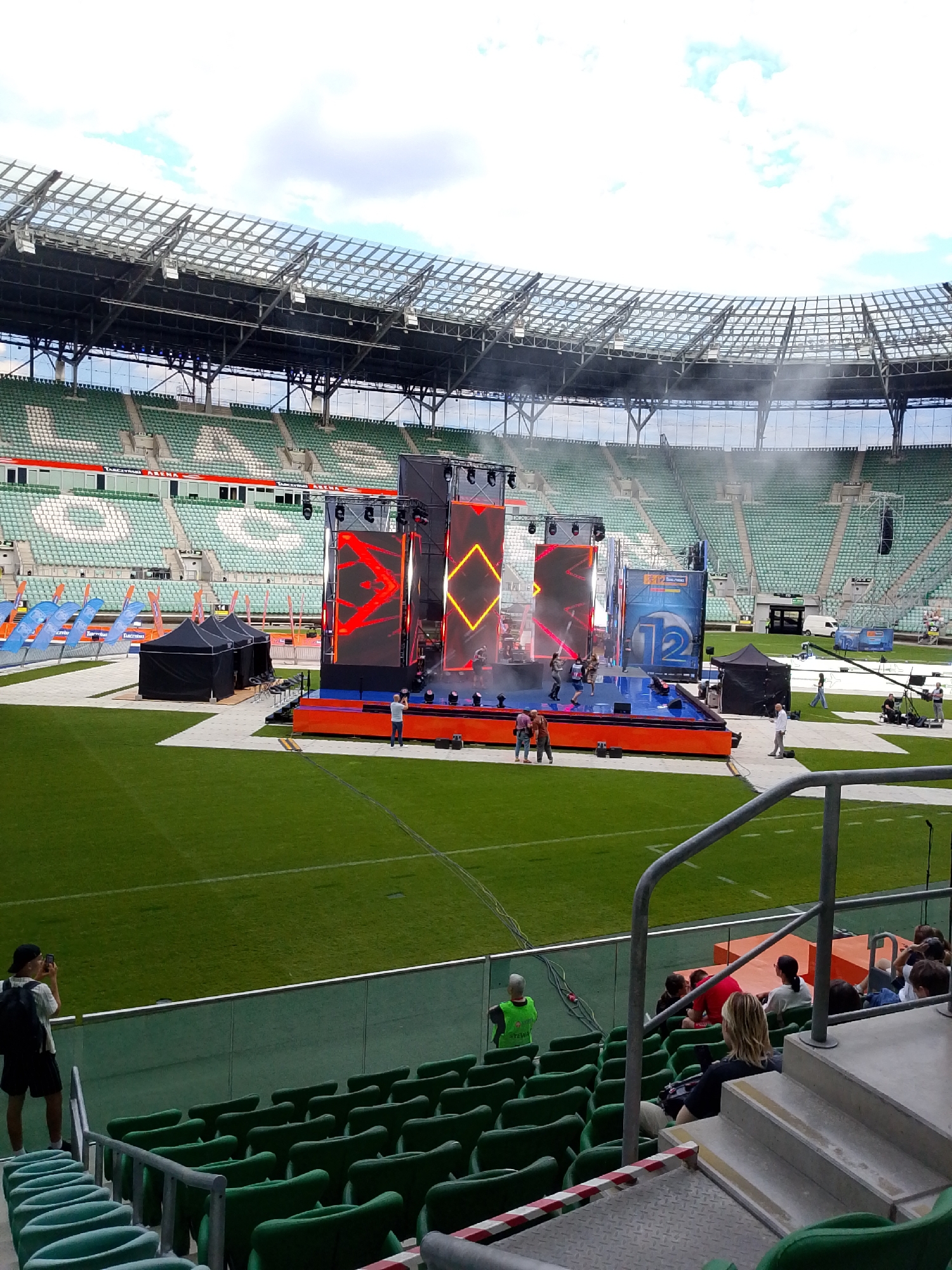 This image captures an outdoor event setup or rehearsal within the Tarczyński Arena in Wrocław, Poland. The scene is dominated by a large, elevated stage positioned on the green grass playing field. The stage features three prominent vertical LED screens displaying dynamic red and orange abstract geometric patterns, with additional lighting fixtures and truss structures. Several individuals, appearing to be performers or crew members in casual attire, are visible on the stage, with smoke or haze effects emanating from the area. To the right of the main screens, a tall blue structure featuring the number "12" and some white text is integrated into the stage design.
Around the stage, on light-colored pathways laid across the grass, several crew members are present. Two black pop-up tents are located to the left of the stage, and another one is to the right. Various cables are visible running across the field. In the mid-ground, rows of empty green and white stadium seats spell out "TARCZYŃSKI ARENA." The stadium's modern roof, constructed with glass panels and metal trusses, is visible against a partly cloudy sky.
In the foreground, from an elevated seating perspective, rows of empty green stadium seats are visible. A grey metal railing spans horizontally across the lower portion of the image. On the left, a person wearing a dark backpack and cap stands, taking a photo with a smartphone. On the right, several individuals are seated or standing in the viewing area, including one person with blonde hair seated closer to the railing. A person in a bright green vest, likely event staff, is also visible in the foreground seating area. Concrete steps with a metal railing and red-and-white striped barrier tape lead down into the seating area from the right.