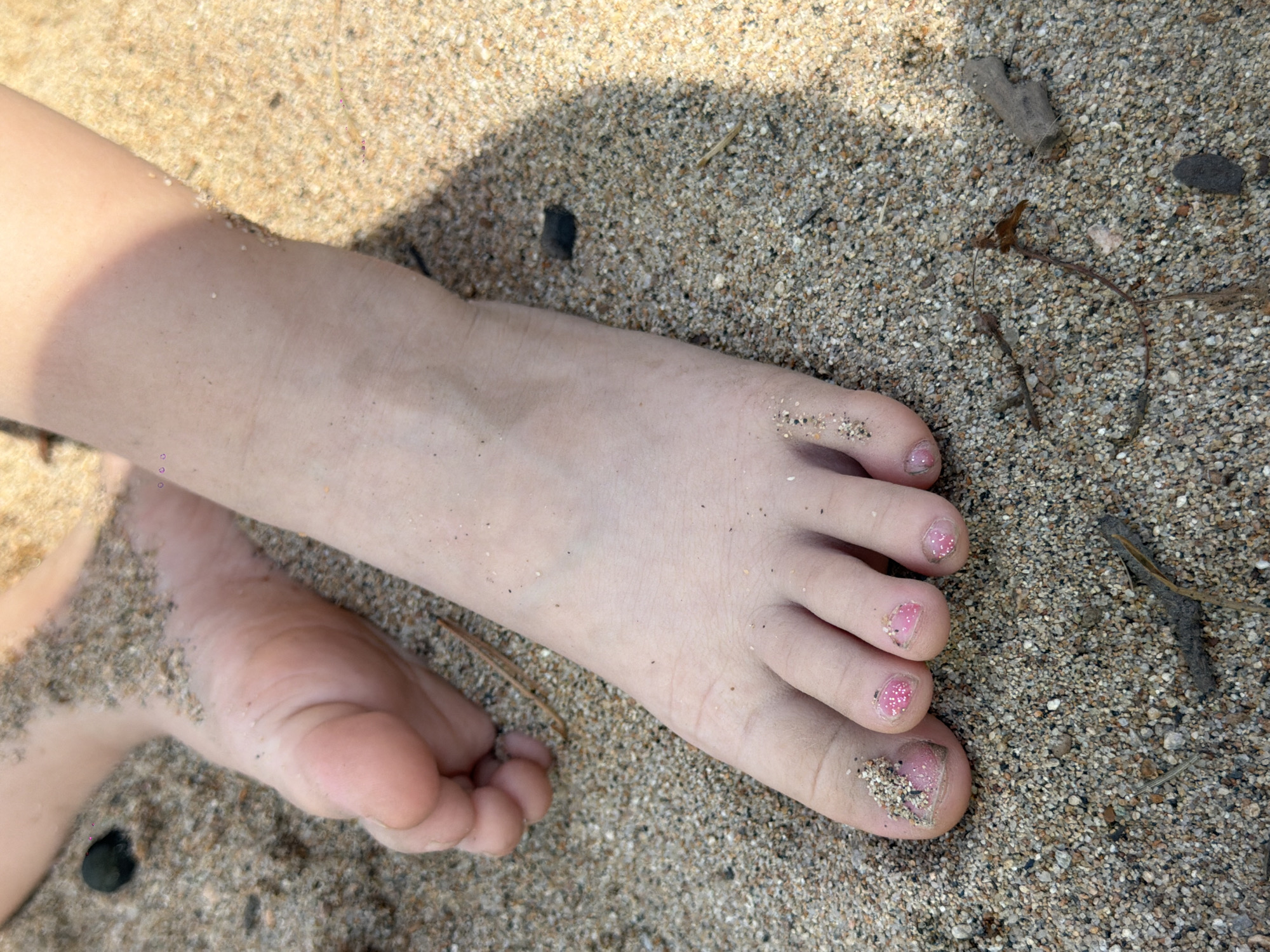 The image shows a close-up view of a child's feet partially buried in sand. The sand is coarse and granular, with small pebbles and twigs mixed in. One foot is more prominent, with the toes visible and painted with pink glitter nail polish. The other foot is partially visible in the background. The lighting suggests it is a sunny day, with a shadow cast across the sand from the right side of the frame. The scene is a beach or sandy area, which could be in Maksimiha, Russia, given the context. The focus is on the feet and the sand, with no other identifiable elements of the surrounding environment.