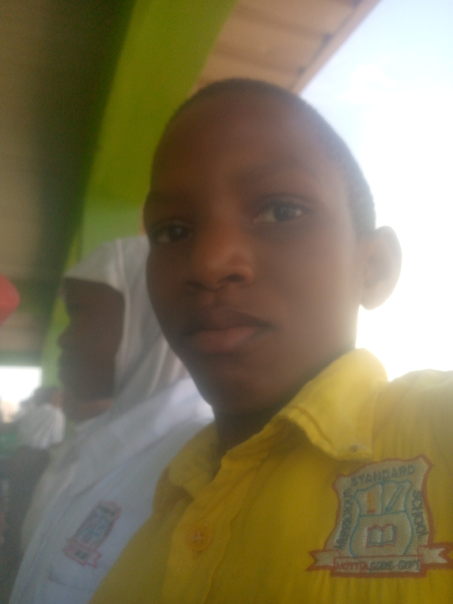 A close-up shot depicts a young person wearing a yellow short-sleeved uniform shirt with a school emblem on the right side. The emblem features the word "STANDARD" and an open book. Another person wearing a white shirt is partially visible in the background to the left. The location appears to be outdoors, possibly in a city with an unknown name. The environment is obscured, but the presence of a green object suggests a natural or possibly an artificial structure in the vicinity.
