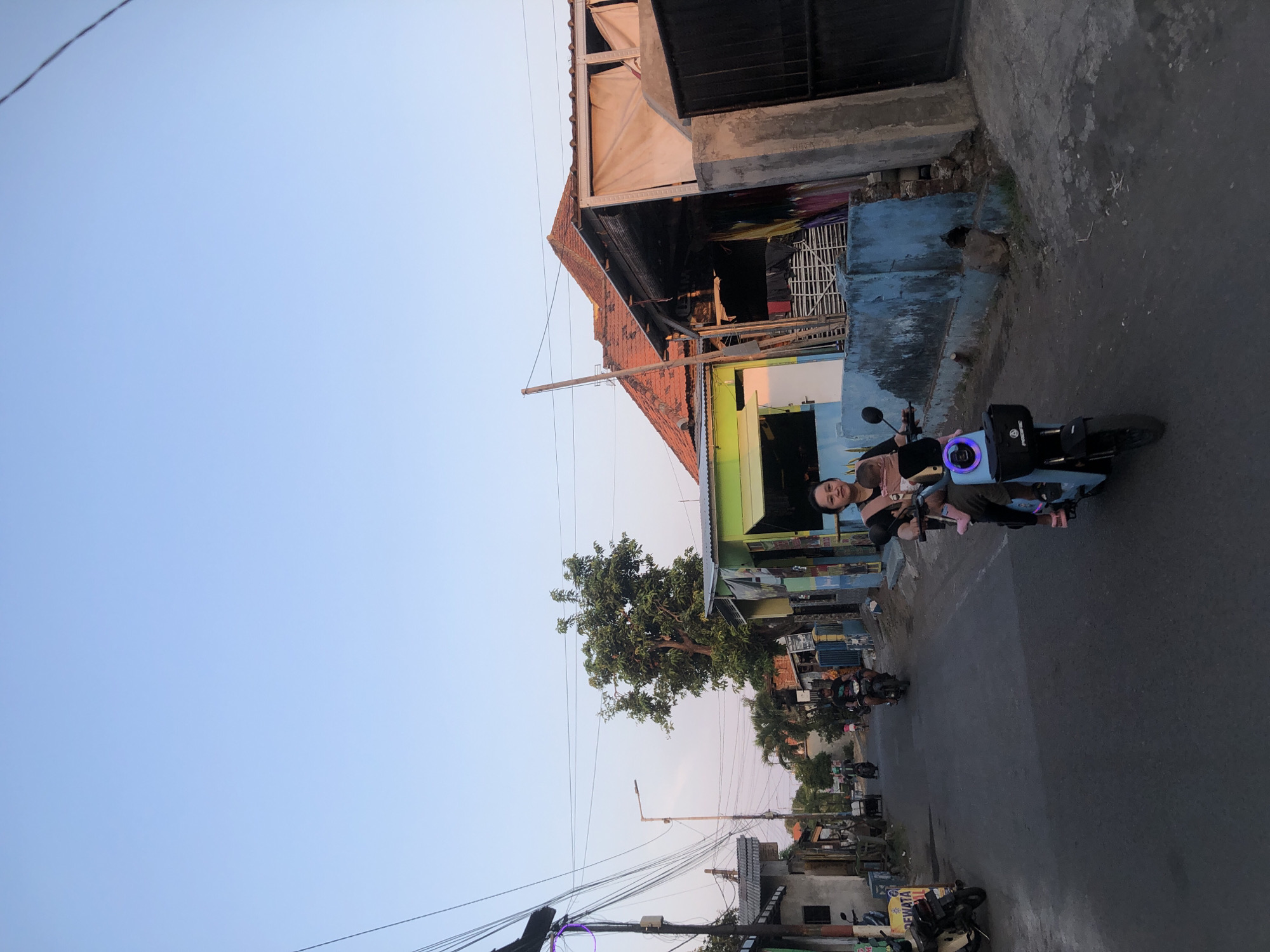 The image depicts a street scene in Tenggulunan, Indonesia. In the foreground, a woman rides a light blue electric scooter with a baby strapped to her chest. She is looking towards the camera with a slight smile. The scooter has a black basket on the front with a "Pacific" logo. The woman is wearing a black t-shirt and light-colored pants, and her feet are adorned with pink sandals.
The street is paved and appears to be a residential area with houses and small shops lining both sides. The architecture is varied, with some houses featuring red-tiled roofs and others having corrugated metal roofing. There are numerous electrical wires strung overhead, supported by wooden and concrete poles.
In the background, other people can be seen riding motorcycles further down the street. A tall tree with dense green foliage stands prominently near the center of the image. The sky is clear and light blue, suggesting it is either early morning or late afternoon. The overall atmosphere is that of a typical day in a modest Indonesian town.