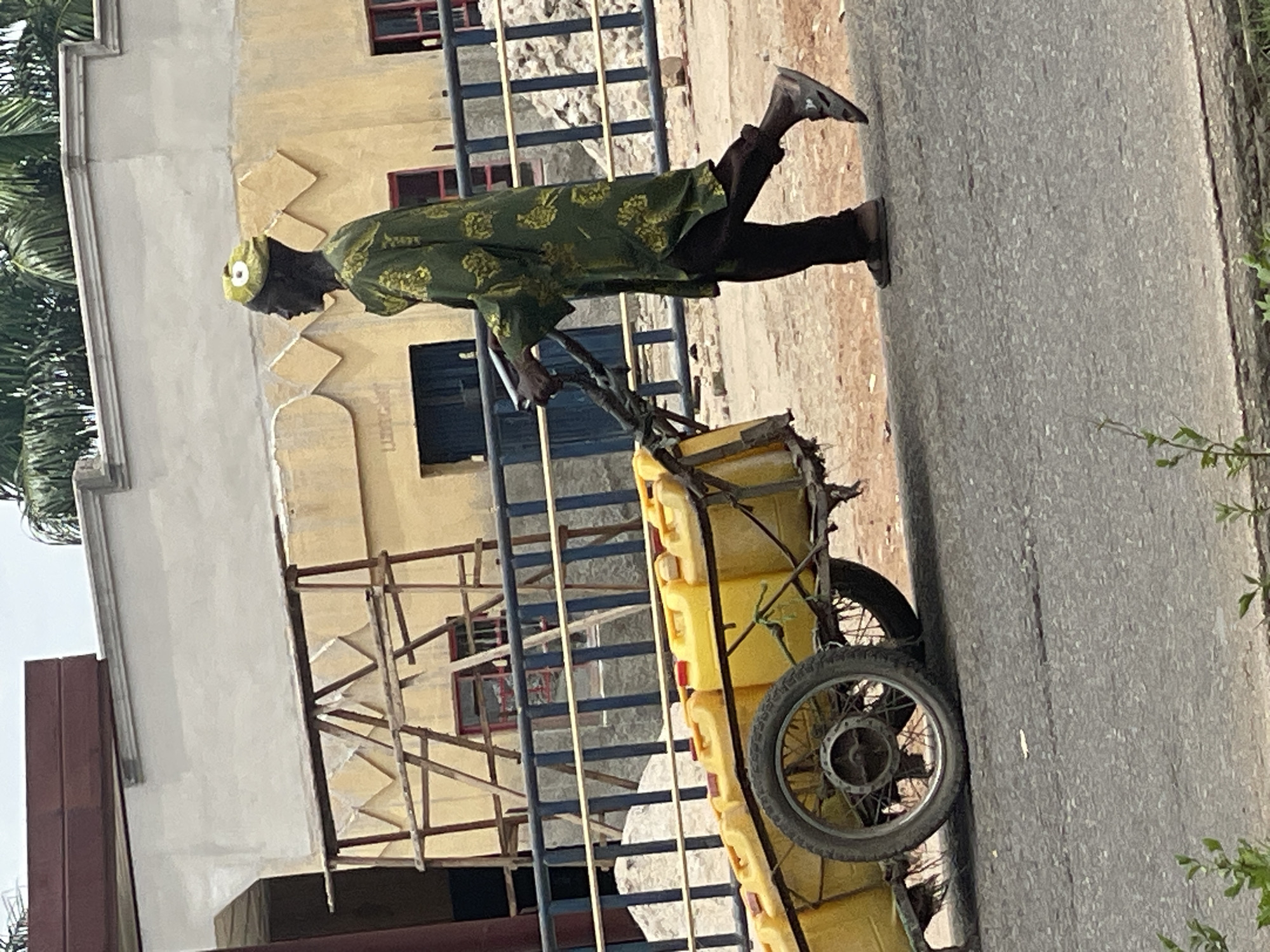 The image captures an outdoor scene, likely in Mararaba, Nigeria, under bright daylight conditions. The main subject is a man of African descent, shown in profile, actively pushing a two-wheeled handcart along a paved road. He is dressed in a vibrant green, patterned long-sleeved tunic, dark trousers, and a light green or yellow cap. He wears sandals on his feet. The man is leaning forward, suggesting effort as he propels the cart.
The handcart is robust, constructed from metal and wood, featuring spoked wheels. It is heavily loaded with at least five bright yellow jerrycans, commonly used for carrying water or other liquids. The road surface is asphalt, and to the left, the ground appears to be sandy or dusty.
In the background, behind a blue metal fence, there are buildings and some foliage. A light yellow building with reddish-brown window frames and subtle decorative diamond patterns is prominent. Next to it, a plainer light grey building is also visible. Wooden scaffolding is erected near the yellow building, suggesting ongoing construction or maintenance. On the far left, green leaves, possibly from palm trees, are visible. The clear, bright lighting indicates it is likely midday or early afternoon. No visible text or discernible emotions are present in the image, which focuses on the man's labor.