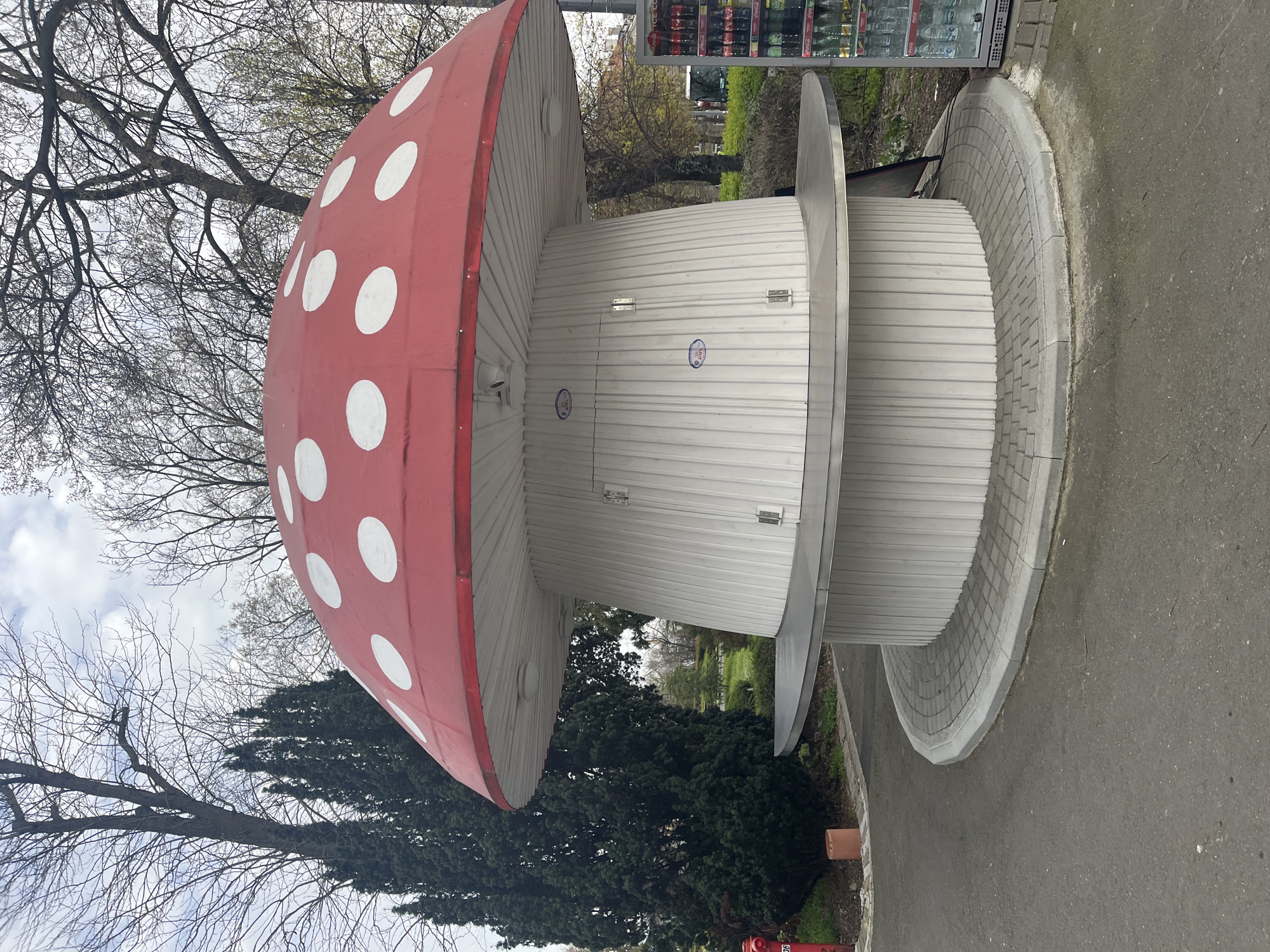 The image depicts a large mushroom-shaped structure, likely a public toilet, with a red cap adorned with white spots. The stem is white with horizontal panels. There are two levels on the mushroom stem. The structure sits on a concrete base with a decorative brick edge. The surrounding environment includes trees, grass, and a paved area. A soda vending machine is visible in the background. The location is likely the Sea Garden in Burgas, Bulgaria. No people are visible.