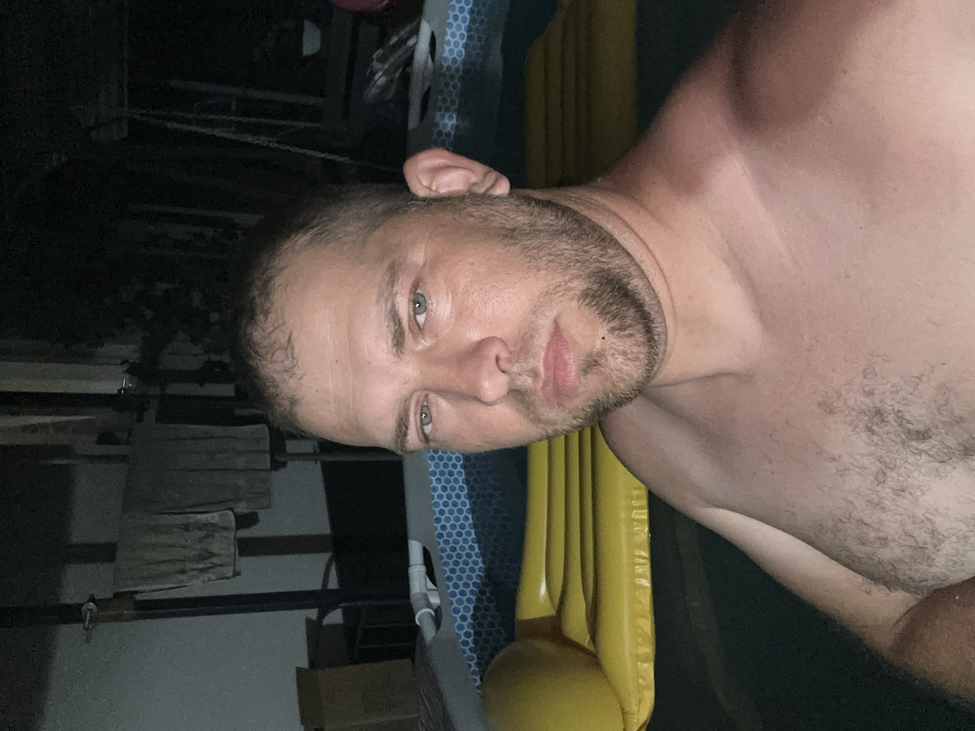 This media file shows a man, shirtless, from the chest up, in what appears to be a dimly lit outdoor setting at night. He has short, dark hair, a beard, and light-colored eyes. The man is positioned in the foreground, with his head tilted to his left. Behind him, to his right, is a large yellow object that resembles an inflatable pool float. To his left and further in the background, there are dark, indistinct structures and what seems to be part of a fence or railing. The scene has a dark and somewhat mysterious ambiance, suggesting it might be late evening or night. No specific activity or event is clearly depicted, beyond the man posing for the camera. There is no visible text in the image. The provided location context is Pîrlița, Moldova.