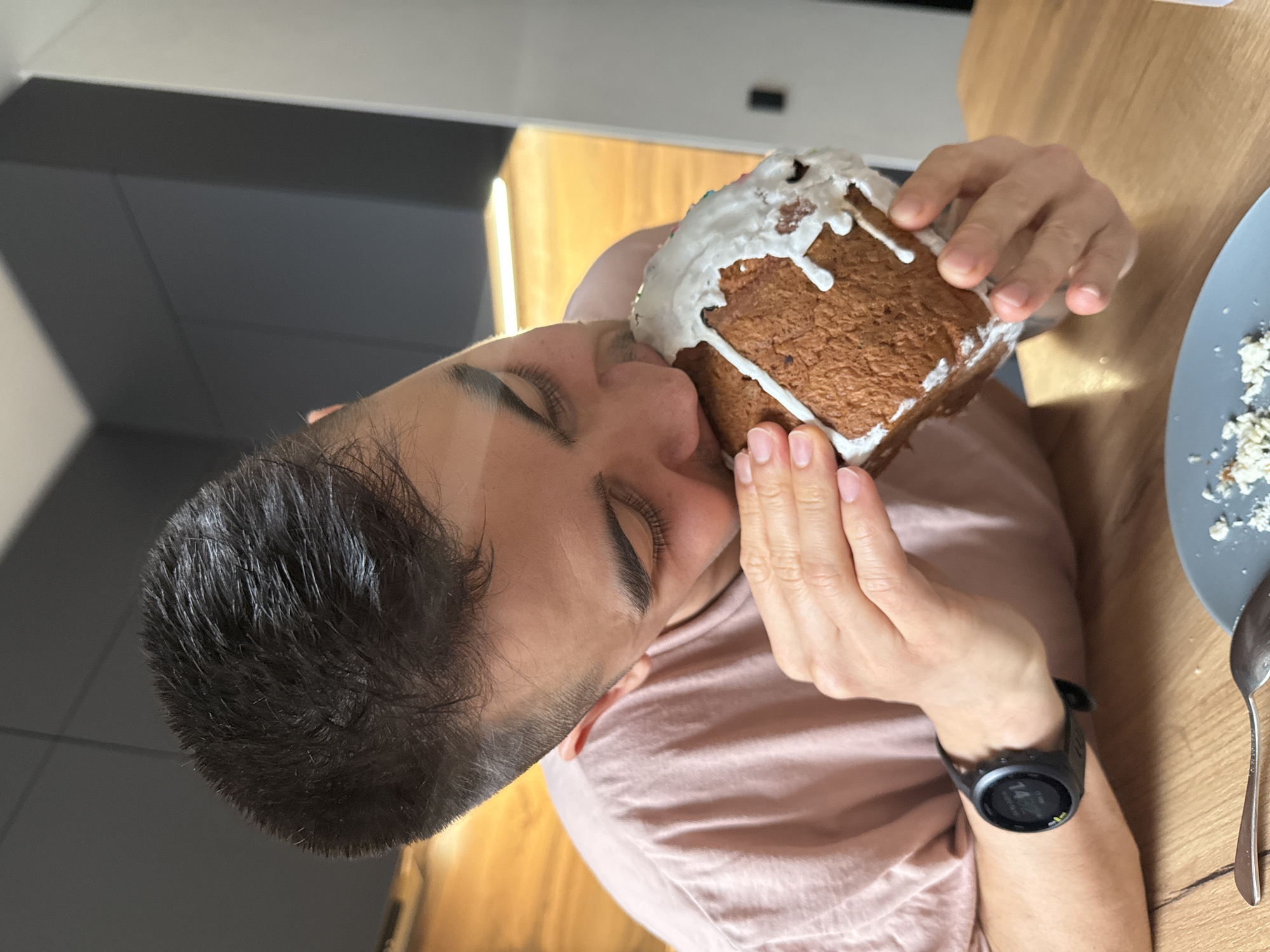 A person with short dark hair and a pink shirt is shown at close range, eyes closed, holding and apparently eating a gingerbread house. The gingerbread house is covered in white icing. A watch is visible on the person's left wrist. Part of a gray plate with crumbs, and a spoon, are visible on a wooden table. In the background are a light-colored cabinet and a dark gray wall. No definitive features of Kazan, Russia are visible.
