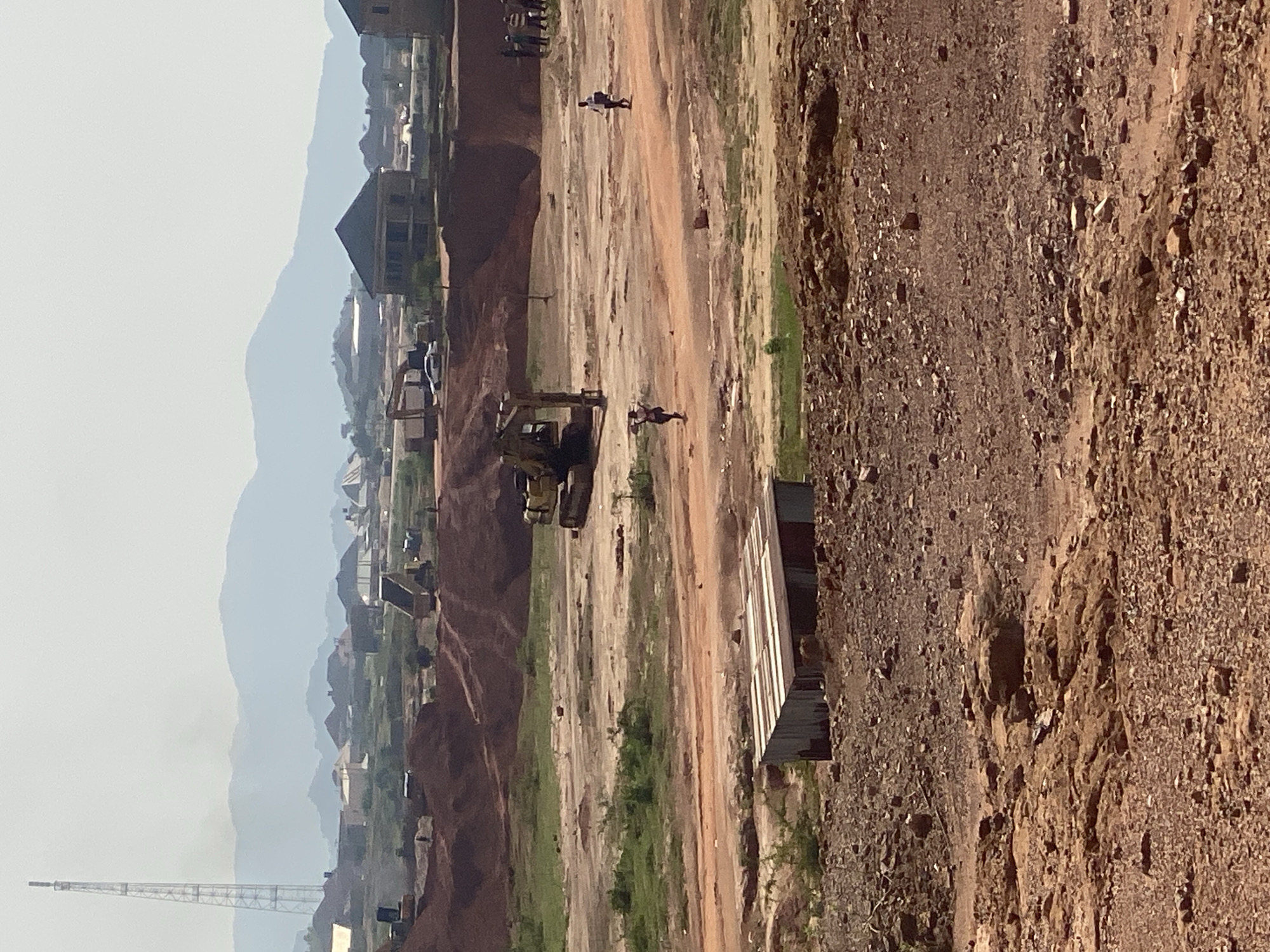 The image captures a construction site in Rayfield, Nigeria, under a hazy sky. In the foreground, a rocky, barren expanse leads to a small, corrugated metal shack with a tin roof. Beyond the shack, a dirt path winds through patches of green grass and exposed earth. Several construction vehicles are visible, including a yellow excavator in the mid-ground and other heavy machinery further in the distance, suggesting ongoing earthmoving operations. A dump truck is also present. Two individuals are walking on the dirt path, one carrying something over their shoulder. In the background, a cluster of houses with pitched roofs indicates a residential area under development. A tall communication tower stands prominently to the left, and distant mountains form the horizon line. The overall impression is one of a developing area with significant construction activity.