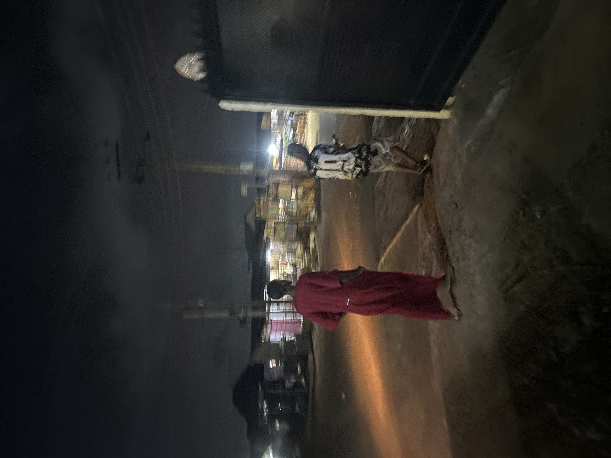 The image depicts an outdoor night scene in an unidentified urban location. The environment consists of a street or sidewalk area, and blurred buildings and streetlights are visible in the background. Two individuals are present: one is wearing a red garment and the other a patterned shirt. The interaction or action is not distinctly depicted due to the image quality and focus.